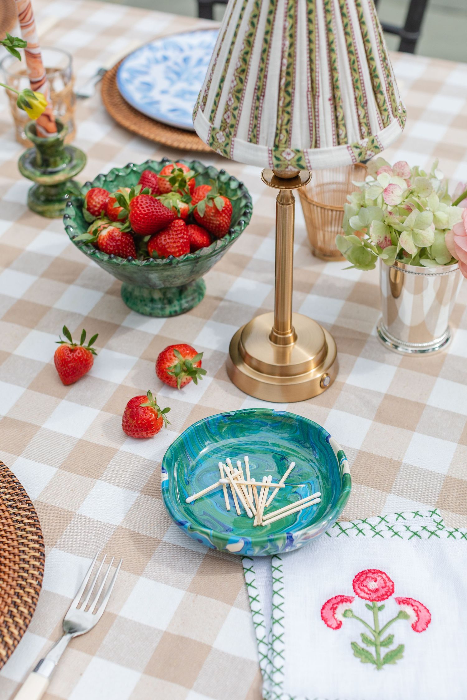 Elegant table setting with gingham cloth, strawberry bowl, brass lamp, toothpick dish, and embroidered napkin.