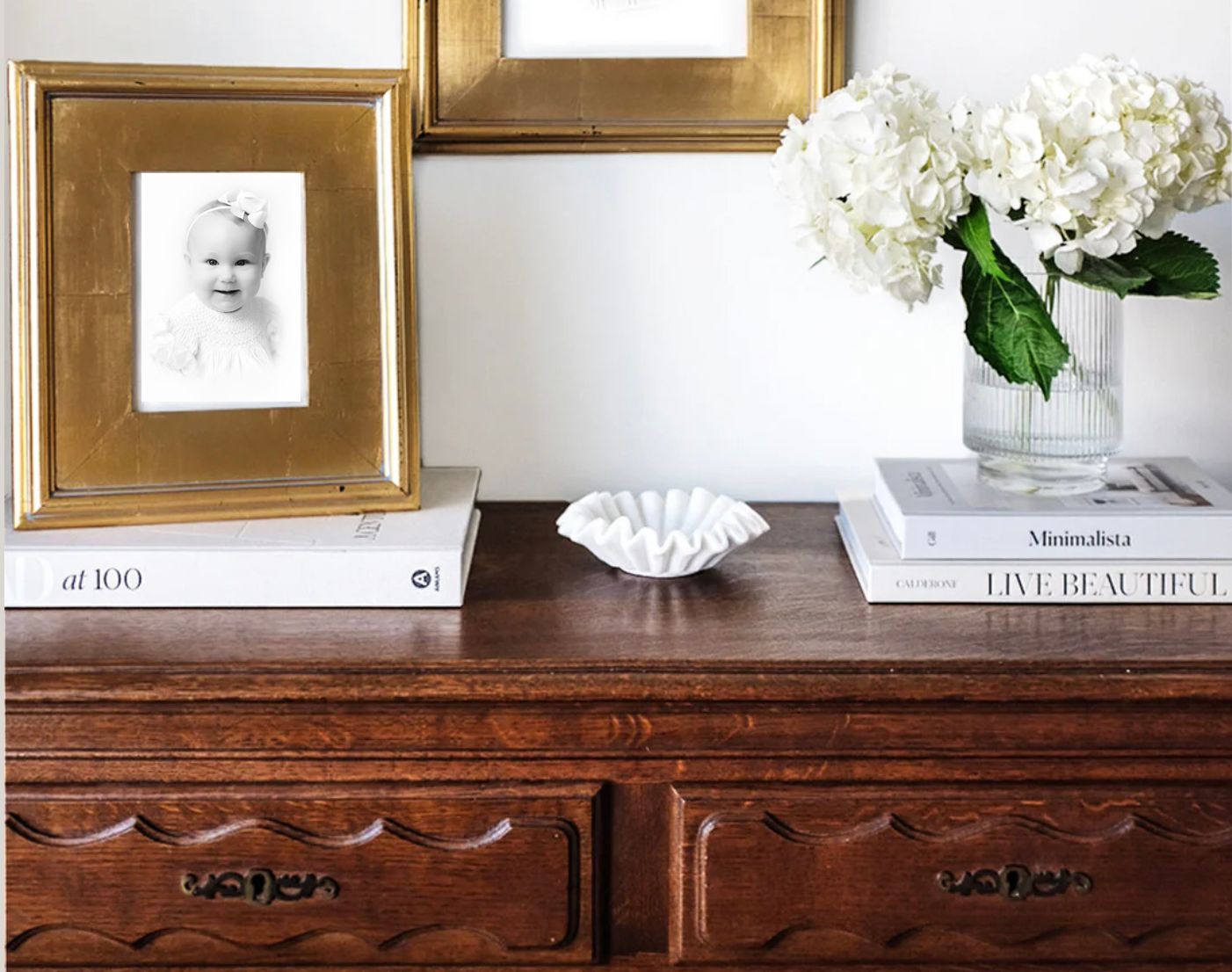 Elegant wooden dresser styled with gold-framed baby photo, white hydrangeas in glass vase, and decorative books.