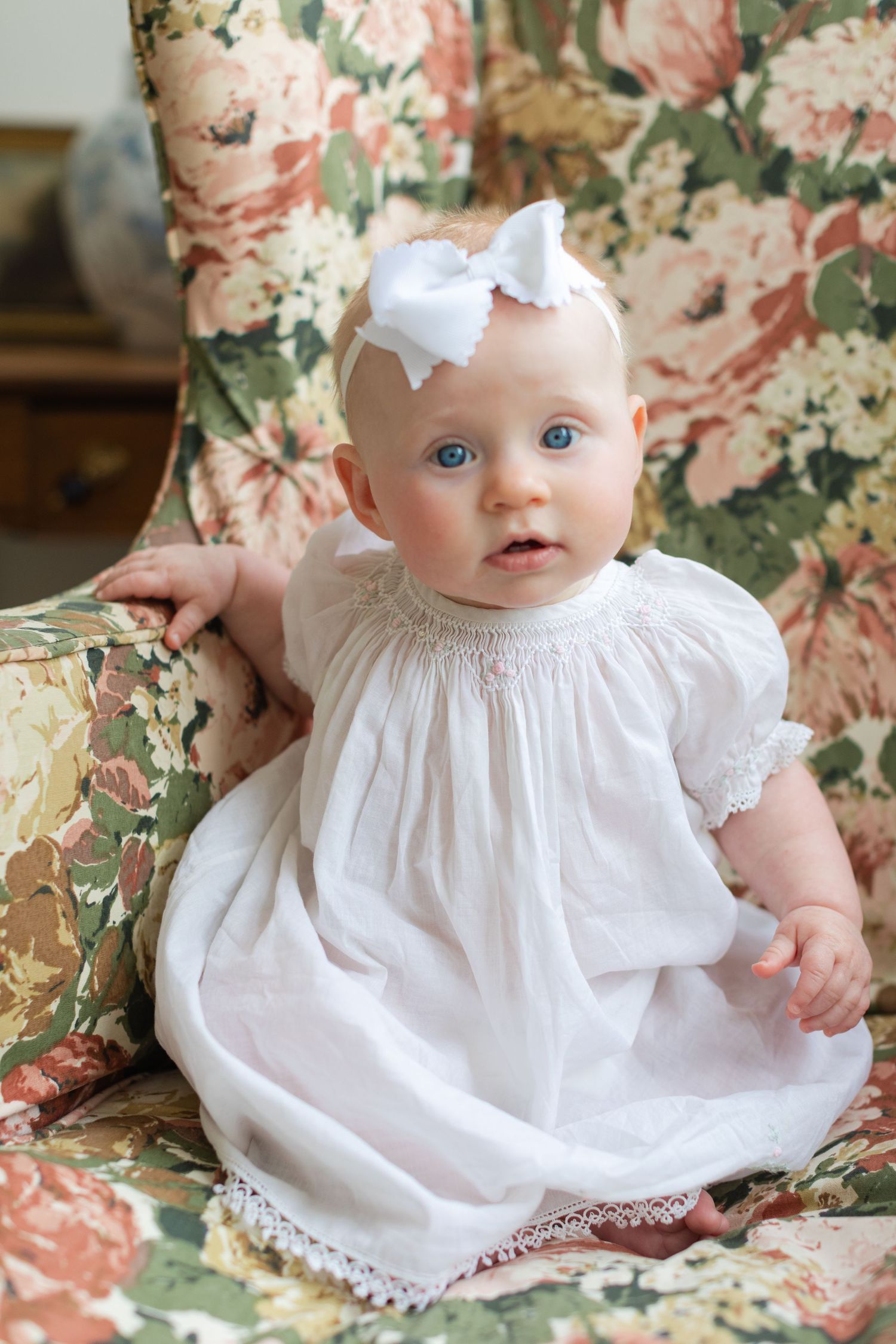 Adorable baby girl in white smocked dress and bow headband sitting in floral armchair with blue eyes.