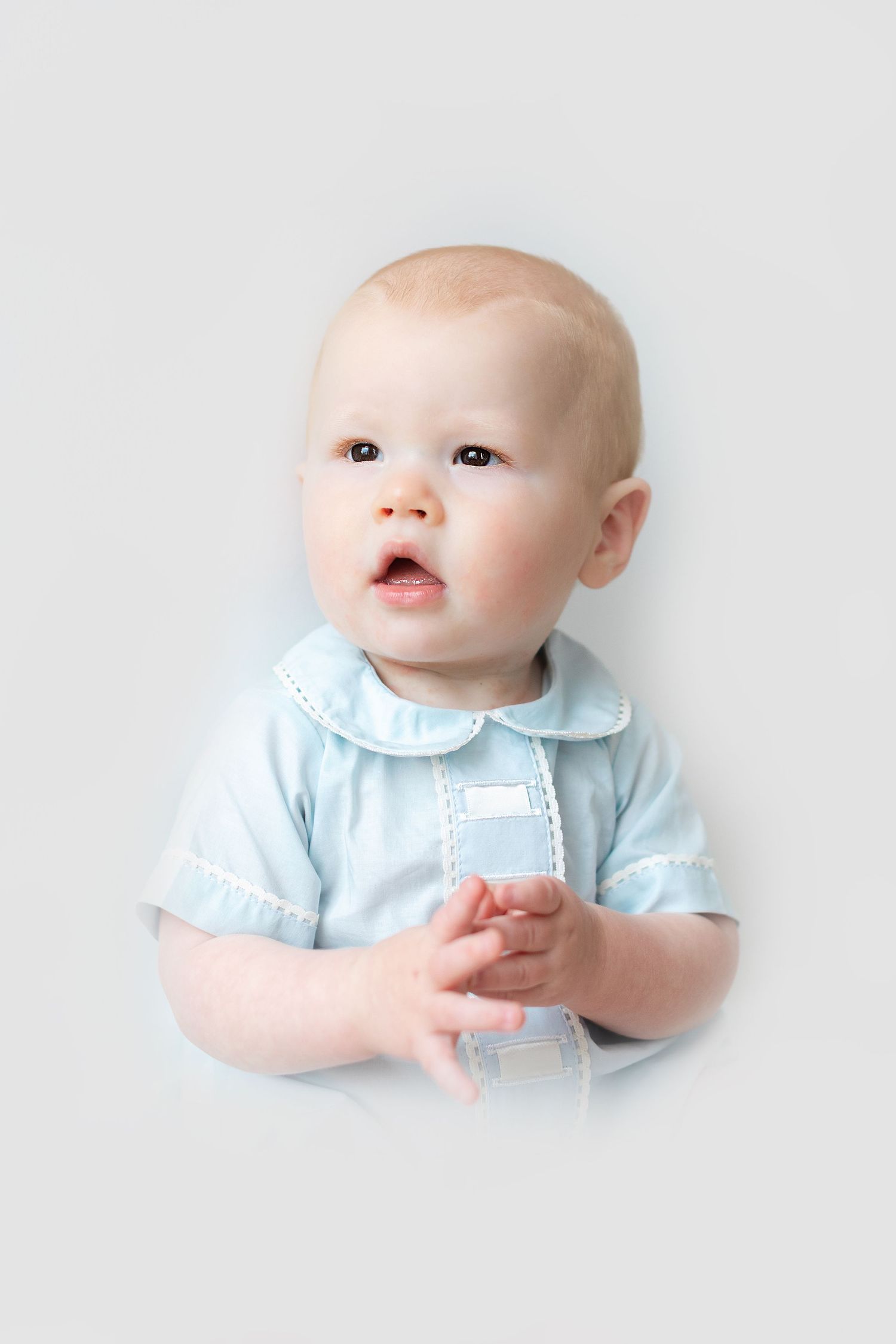 Adorable baby in a light blue outfit with white trim sits against a gray background, looking up curiously.