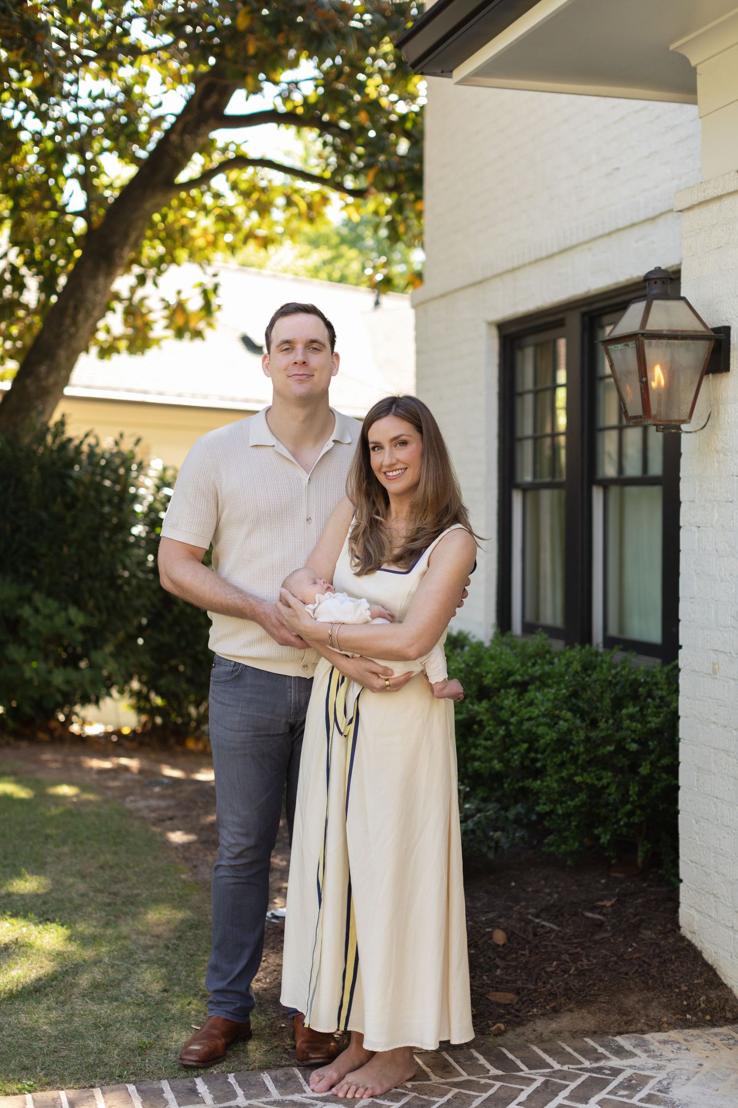 A couple stands outside their home holding a newborn baby, dressed in neutral tones on a brick pathway.