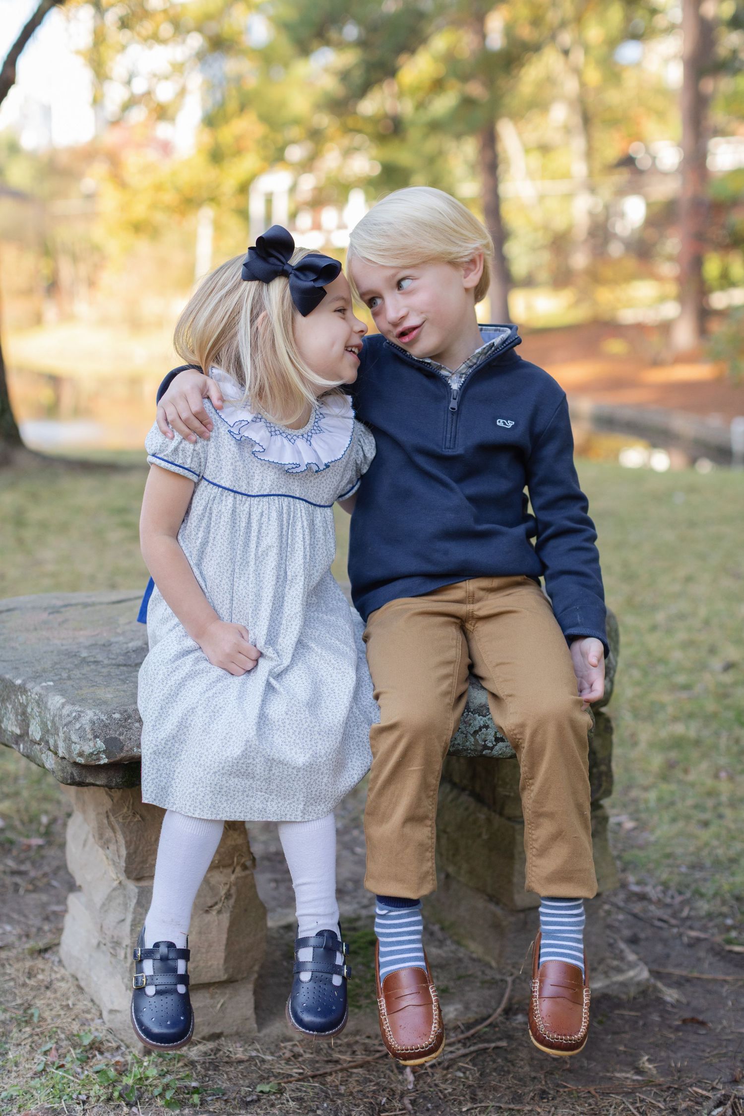 Two children sitting on a wooden bench in an autumn park wearing coordinated navy and khaki outfits.