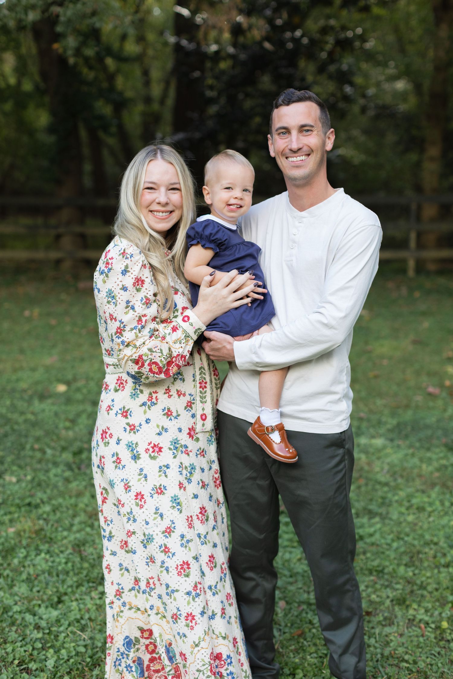 A family poses together outdoors on grass, with a baby wearing navy blue and parents in white and floral print clothing.