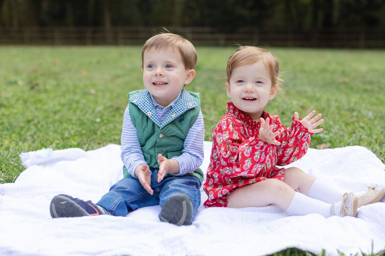 Two young children in casual clothing sit together on a white blanket outdoors on green grass.