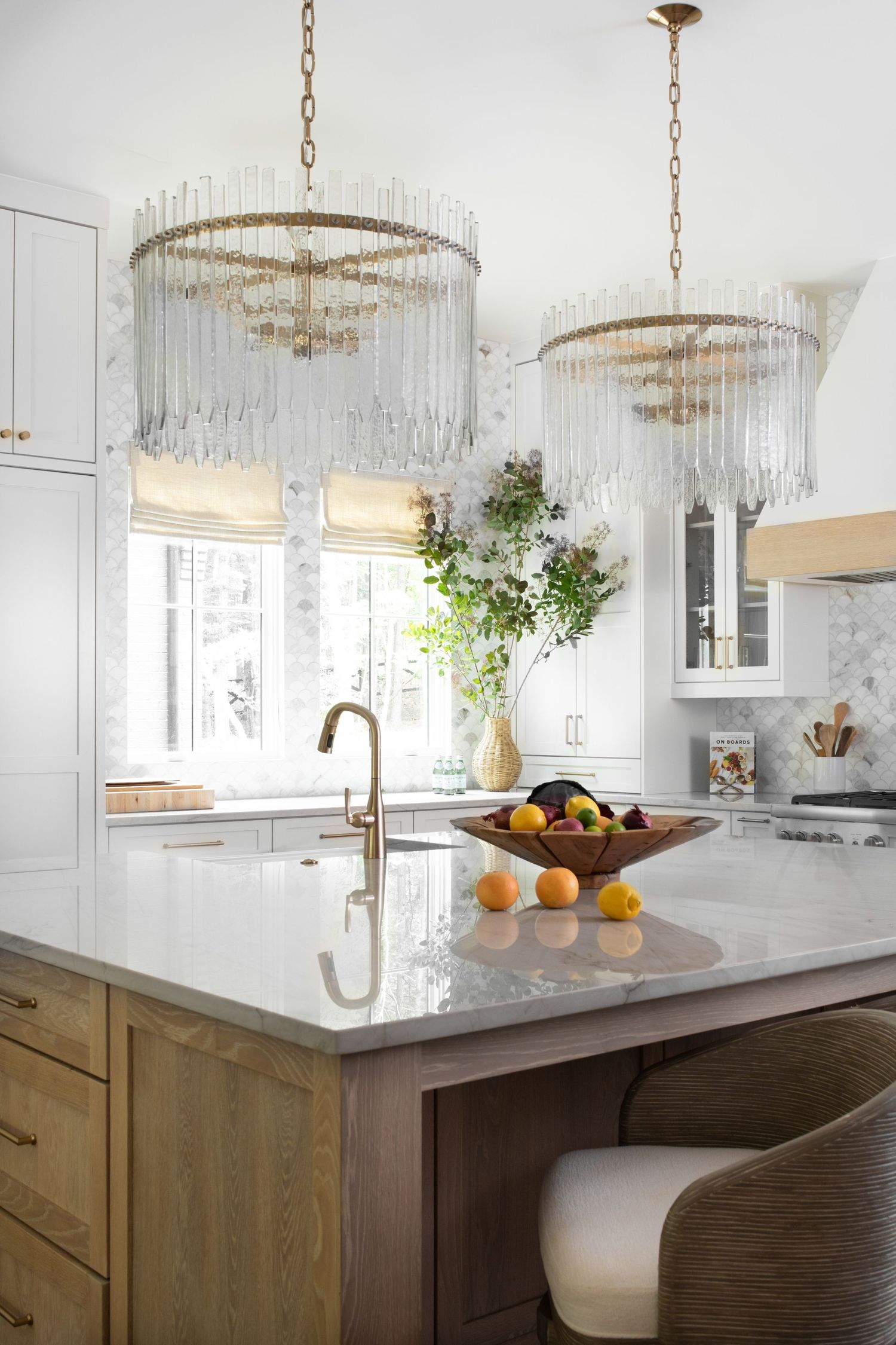 Elegant kitchen with brass crystal chandeliers, marble island, wood cabinetry, and a fruit bowl centerpiece.
