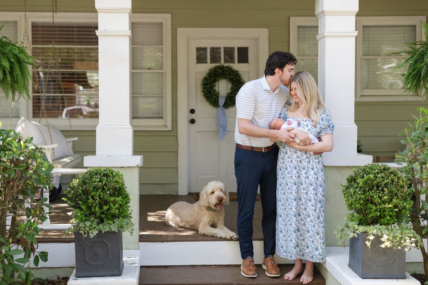 Young family with newborn baby and golden doodle dog posing on charming green home front porch.