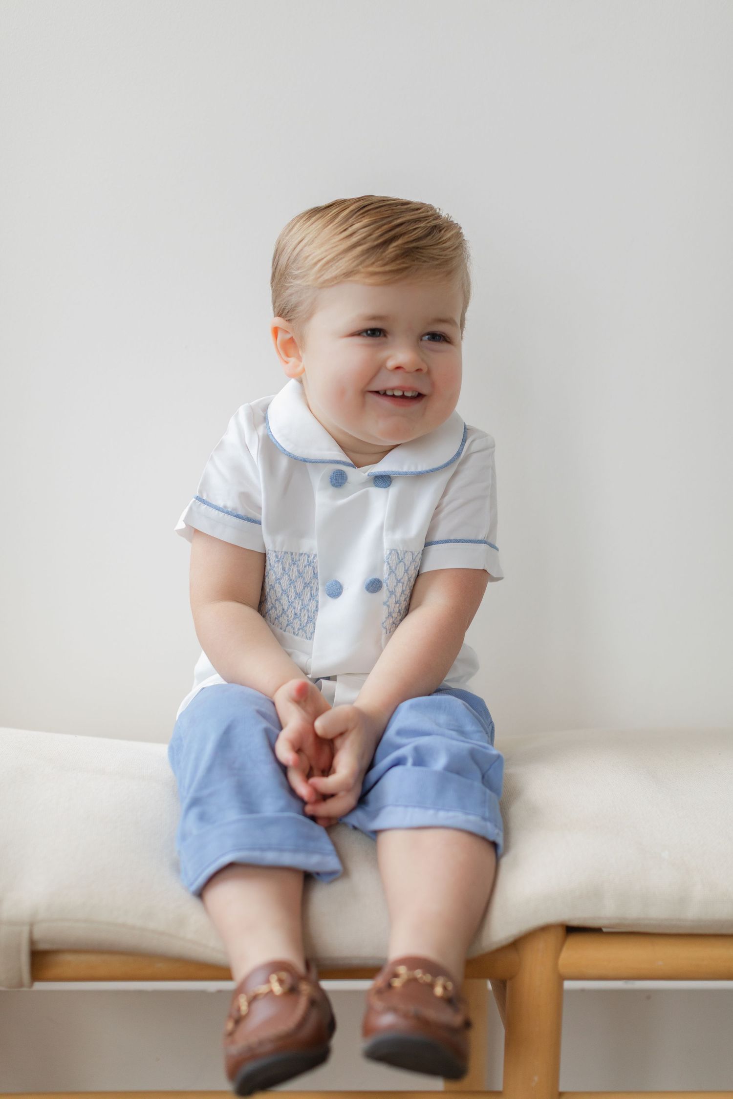 Small child in light blue and white formal outfit with brown leather shoes sits on cushioned bench against white wall.