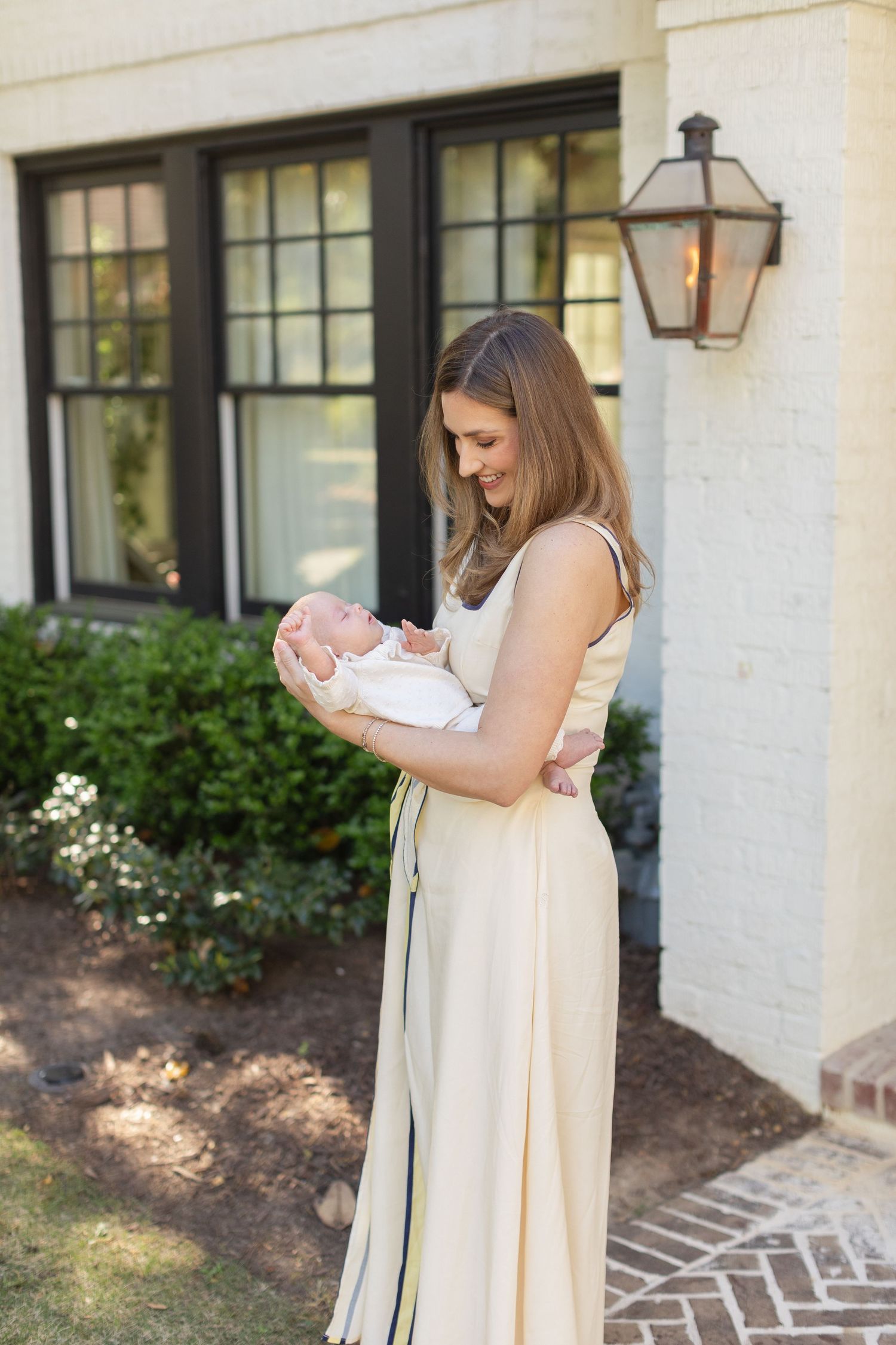 New mother in cream dress tenderly holding her newborn baby outside a brick home.
