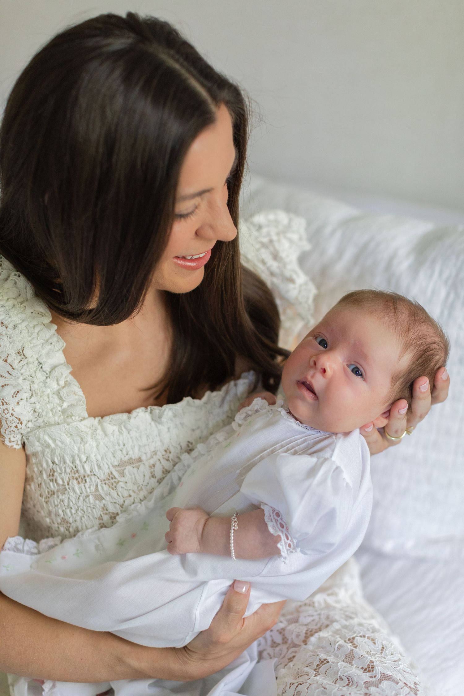A loving mother in white lace holds her newborn baby, both dressed in white, sharing a tender moment.