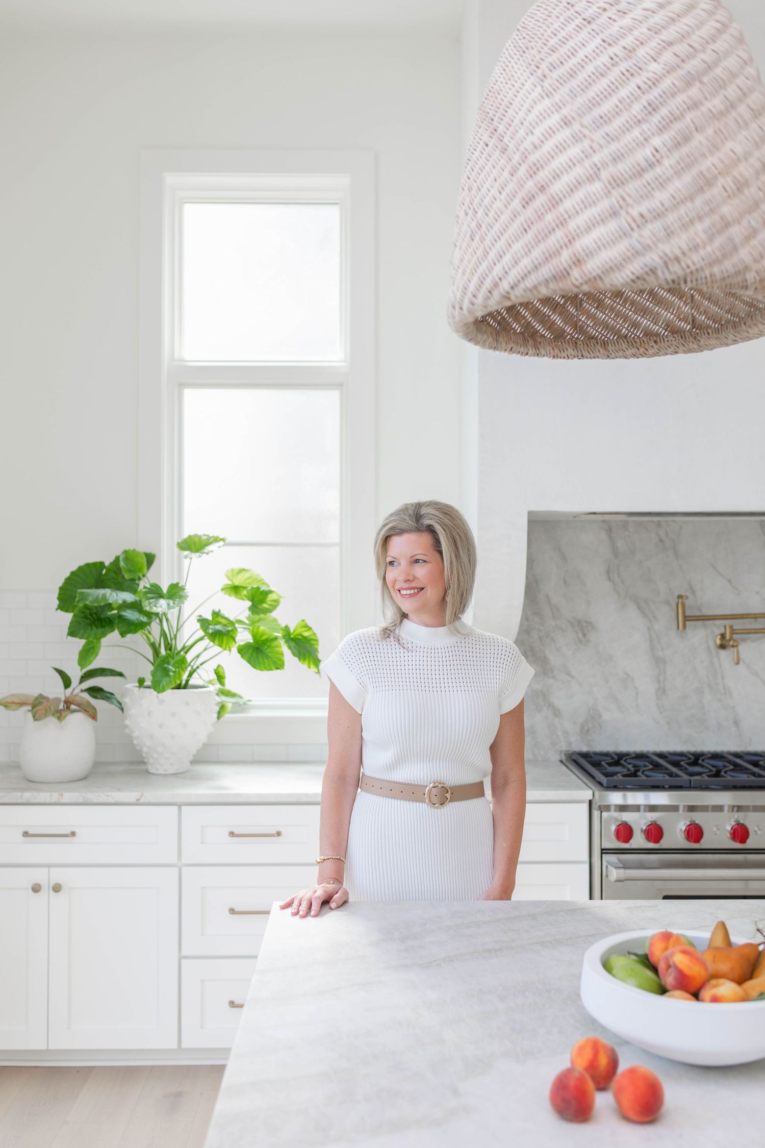 Woman in white dress standing in a bright modern kitchen with marble countertops, white cabinets, and a woven pendant light.