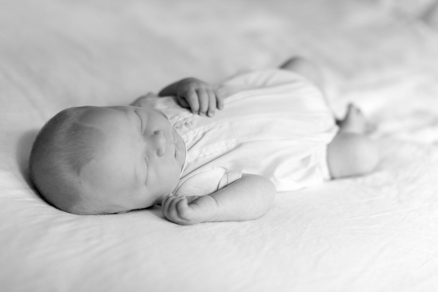 Black and white photo of an infant sleeping peacefully on white bedding.