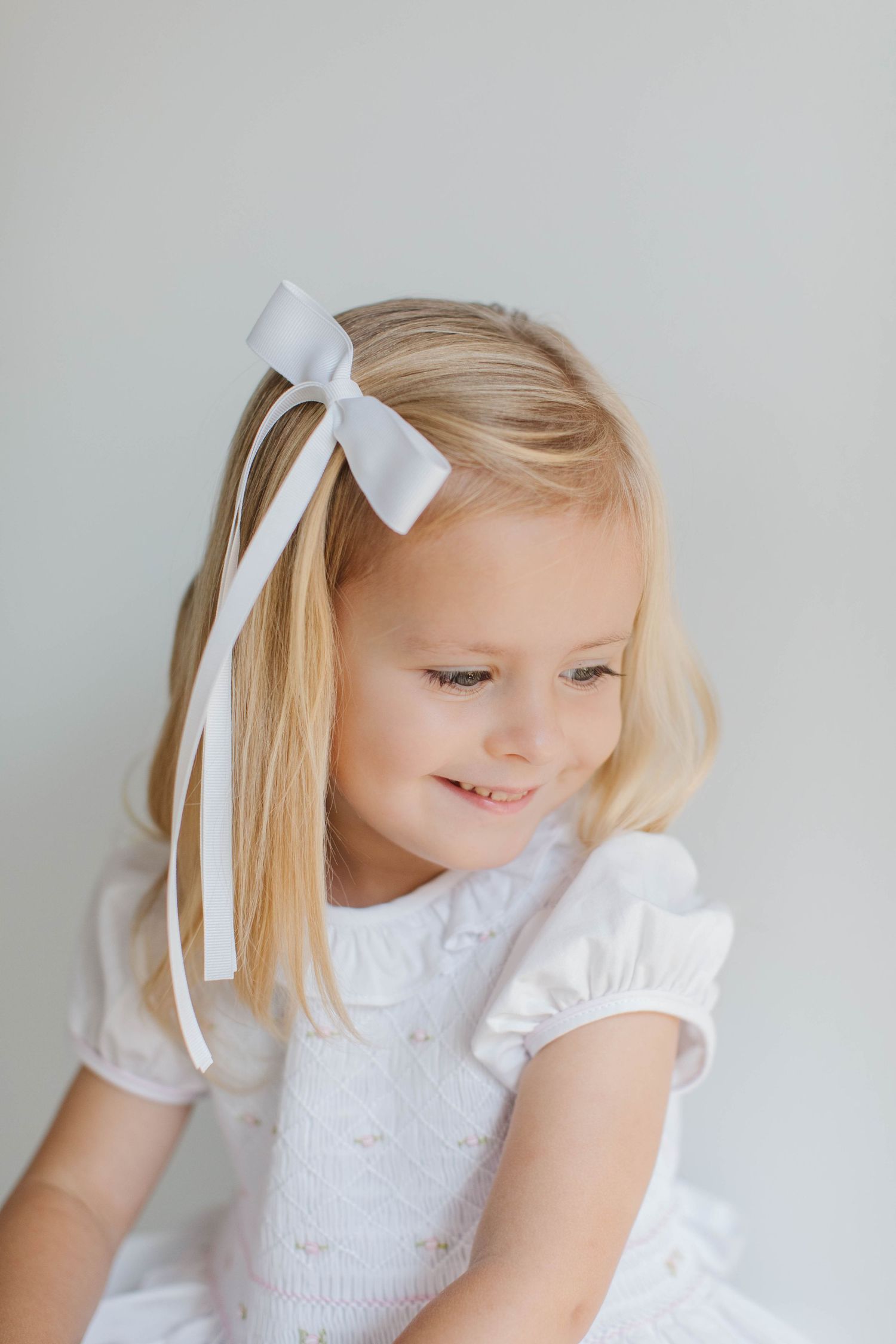 Young child in white dress with white ribbon hair bow smiles sweetly while looking down against light background.