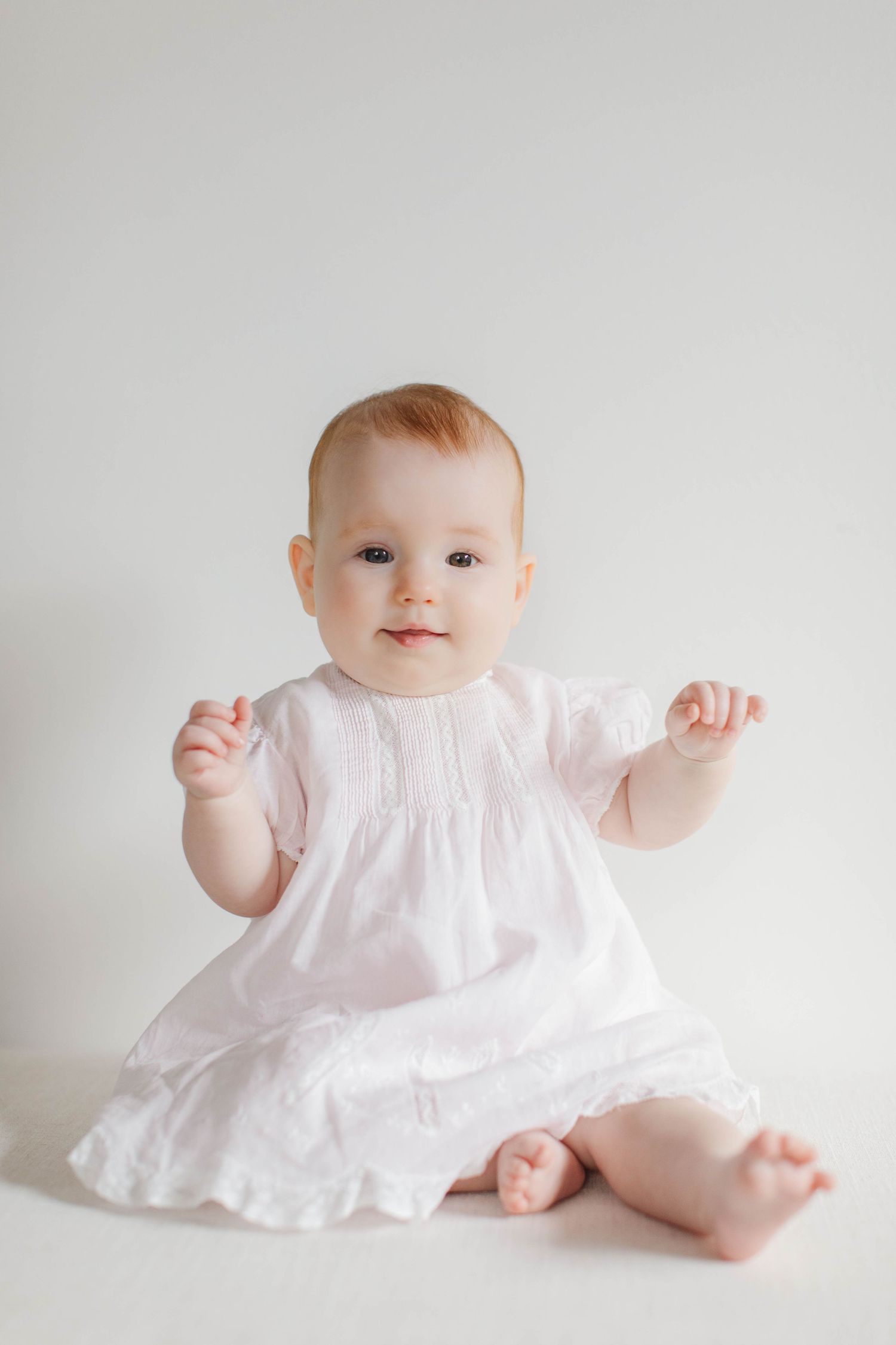 Baby sits in white dress against light background showing playful expressions and movements in studio portraits.