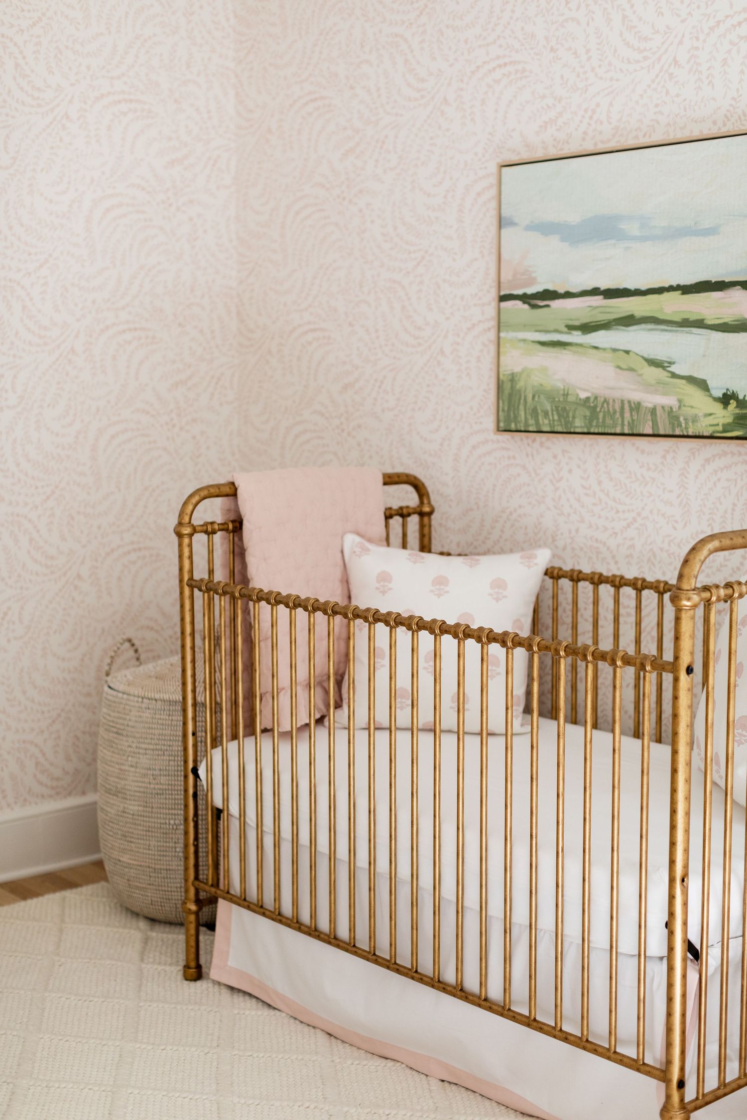 Gold metal crib with pink and white bedding sits beneath a coastal landscape painting in a nursery with textured walls.