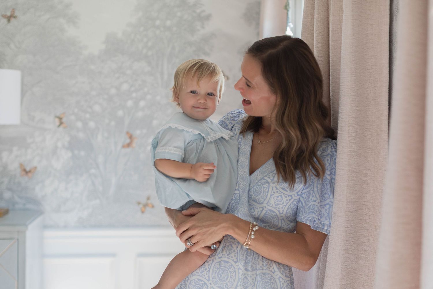 Parent in a floral dress holds a baby wearing a light blue outfit near a window in a well-lit room.