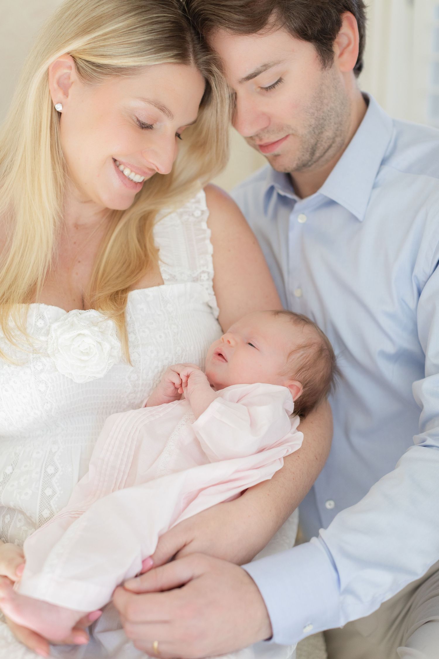 Parents in white and blue outfits share tender moments with their newborn baby wrapped in soft pink blankets.