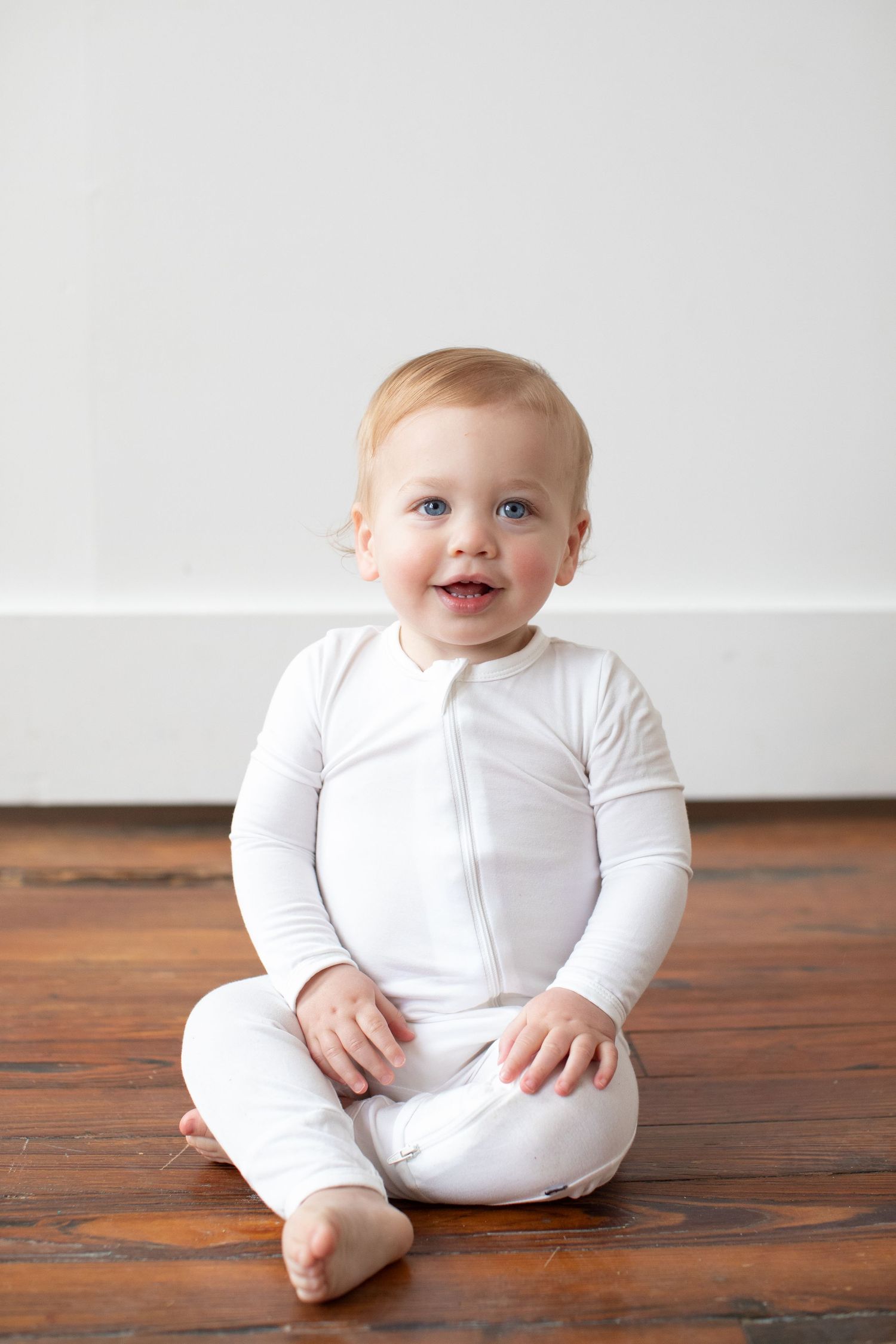 A baby in white clothing sits on a wooden floor in a progression of three similar poses.