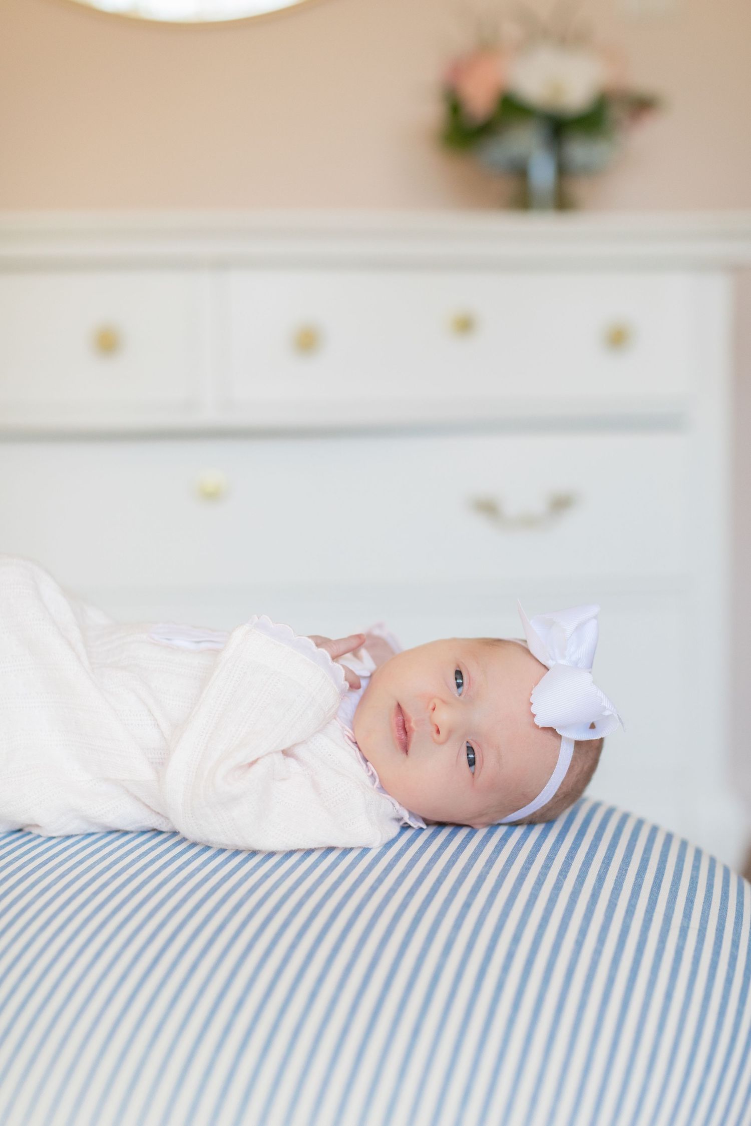 Baby wearing white clothing lies on blue and white striped bedding while looking towards the camera.