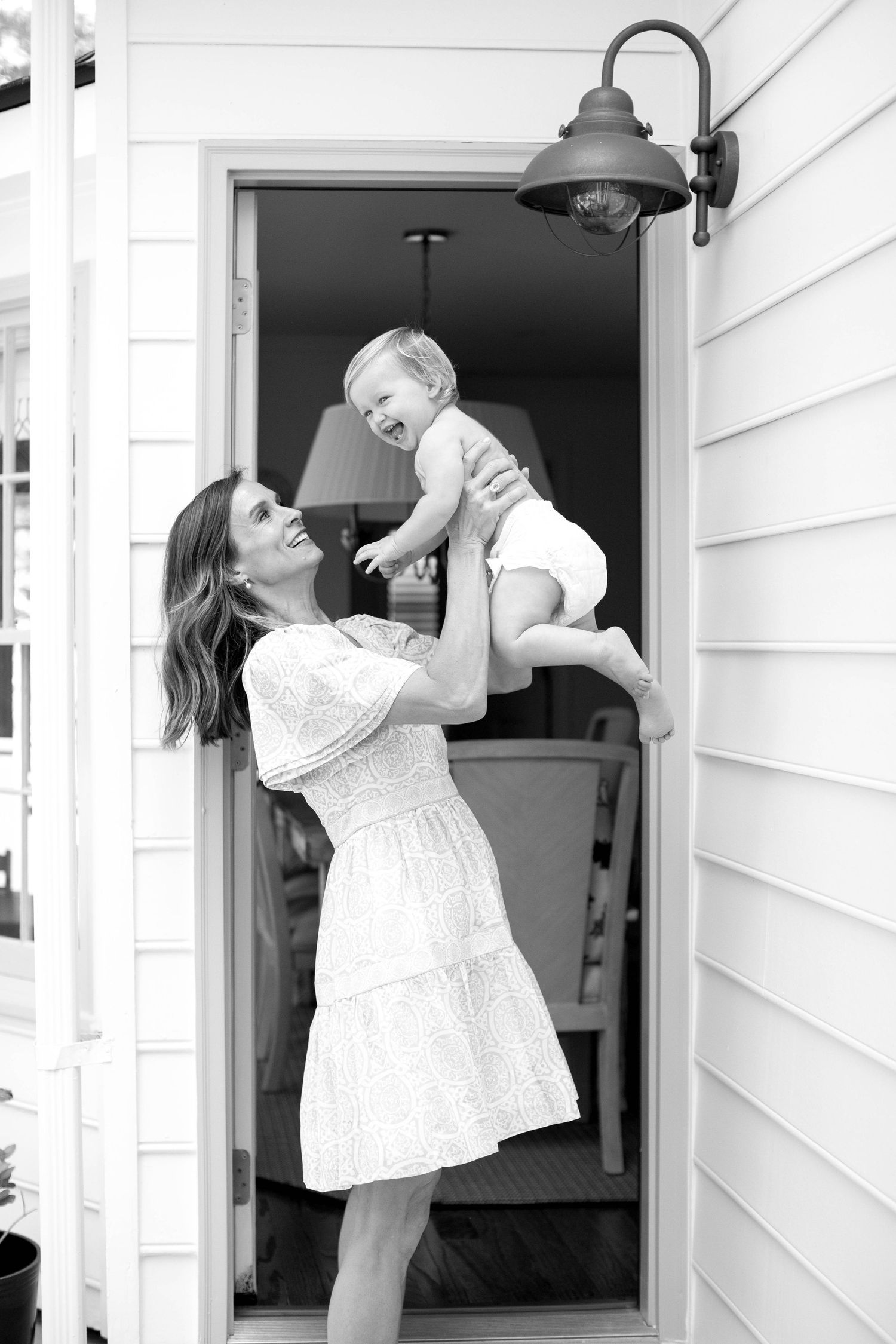 Woman in white lace dress lifts and plays with baby against white exterior wall.