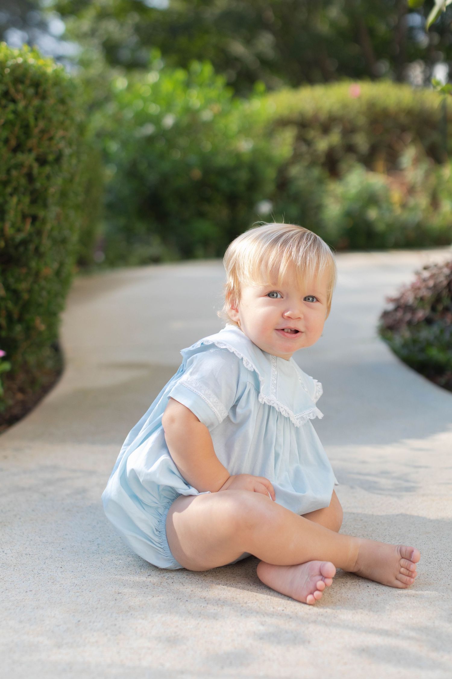 A small child in a light blue dress romper sits on a garden pathway while smiling.