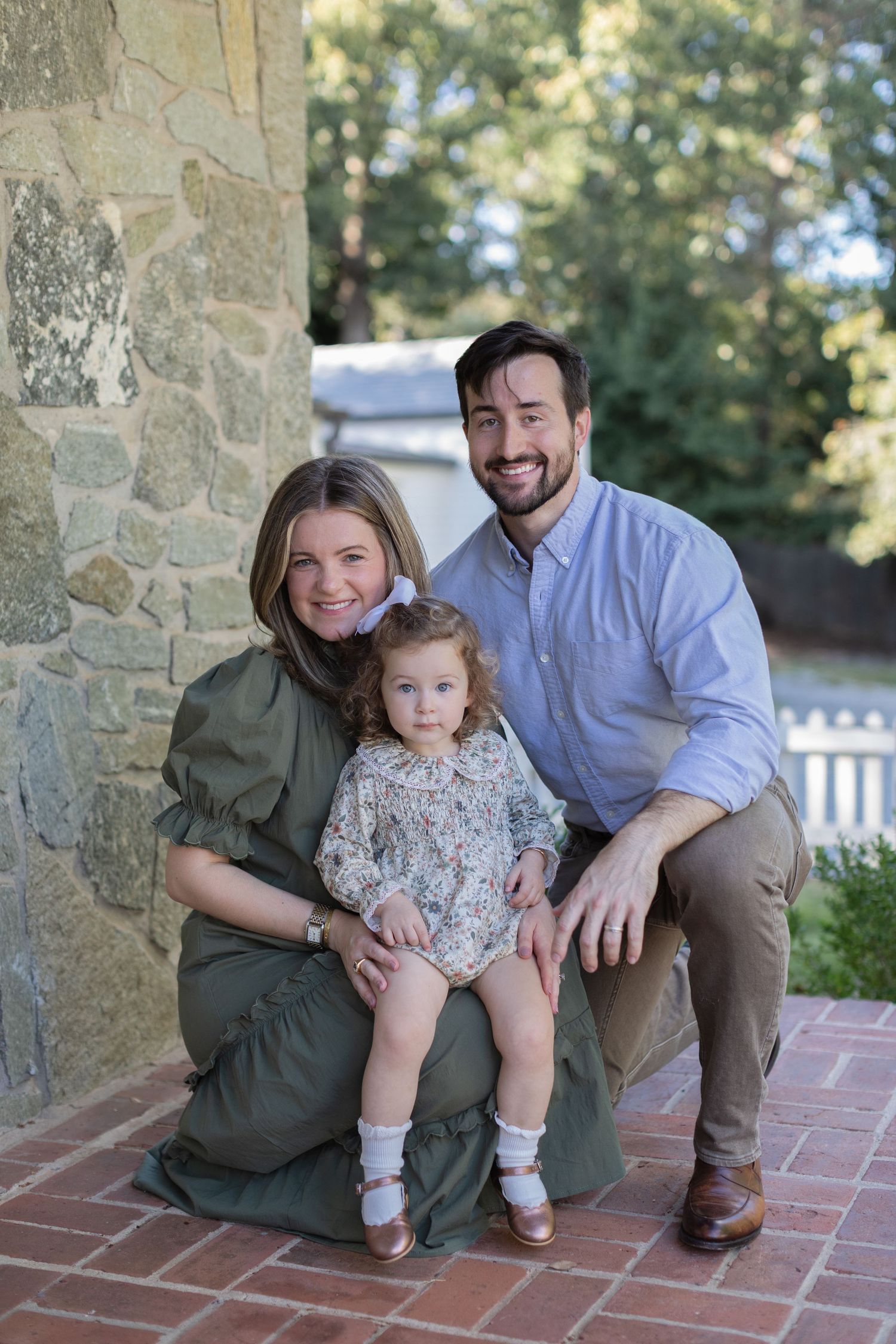 A family poses together on brick steps near a stone wall on a sunny day outdoors.