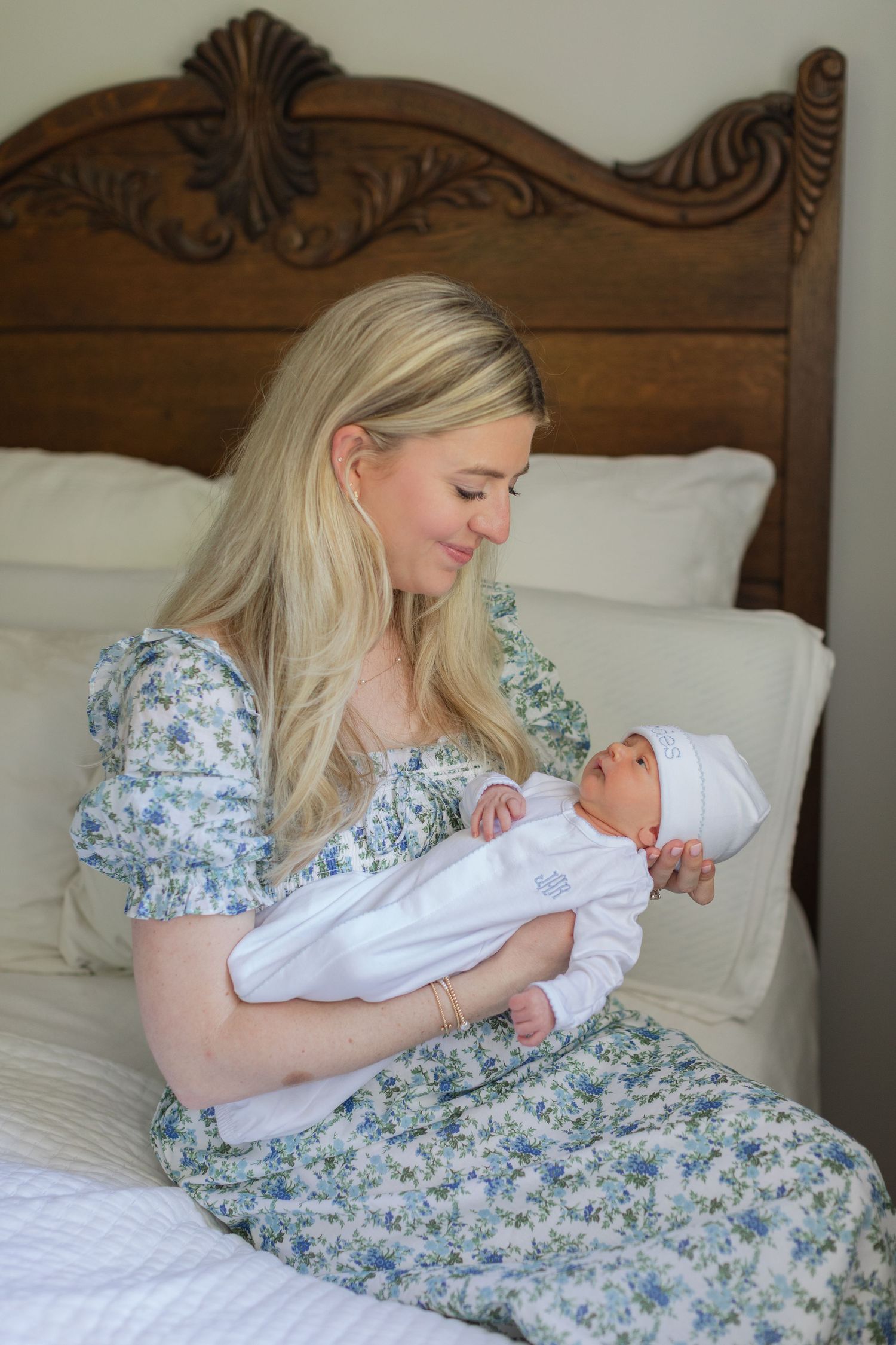 New mother in blue floral dress tenderly holding sleeping newborn baby on vintage wooden bed.