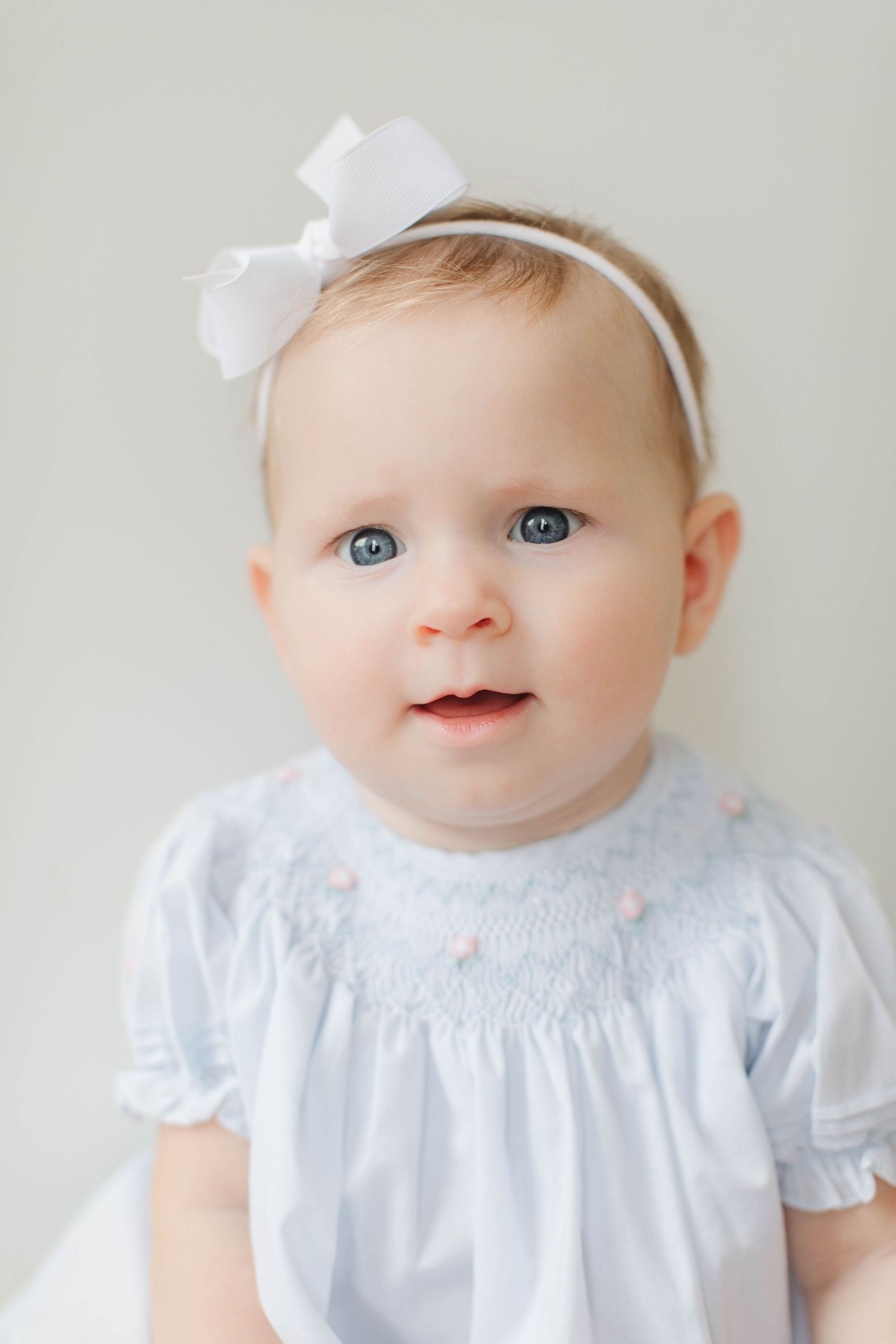 Sweet baby in white smocked dress with white bow headband and bright blue eyes.