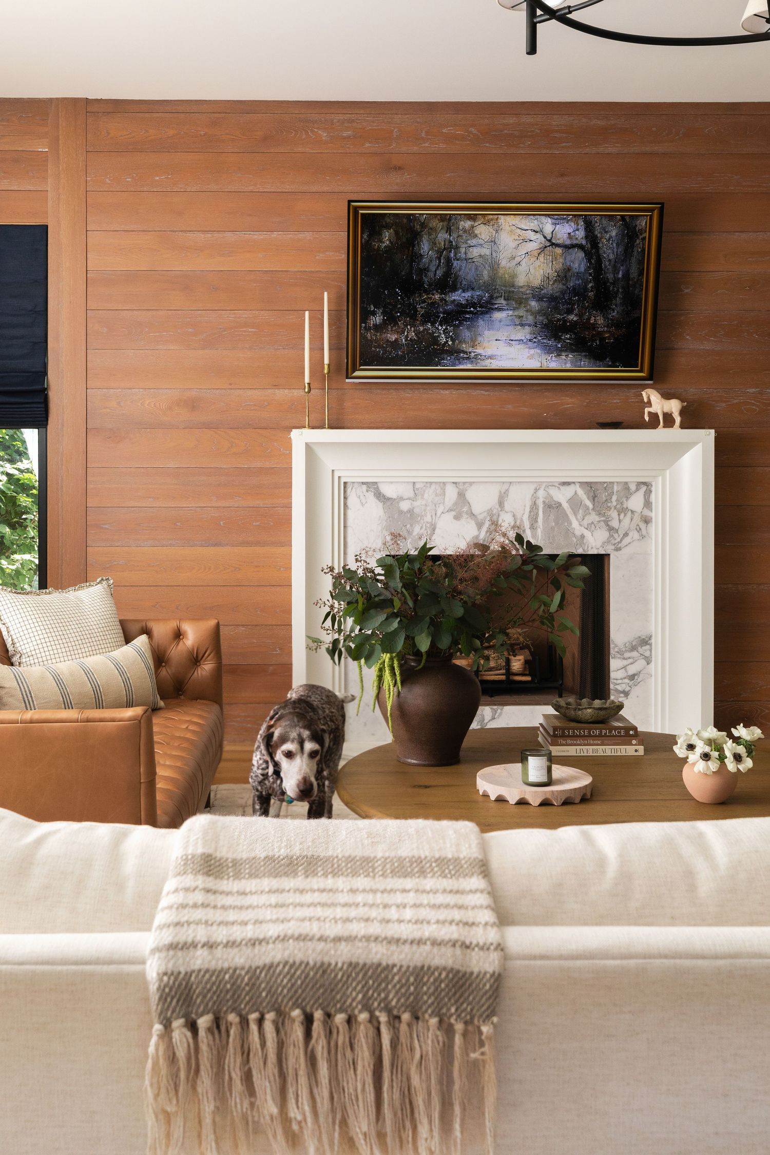 Cozy living room with wood-paneled walls, white marble fireplace, landscape painting, and a dog standing near a coffee table.