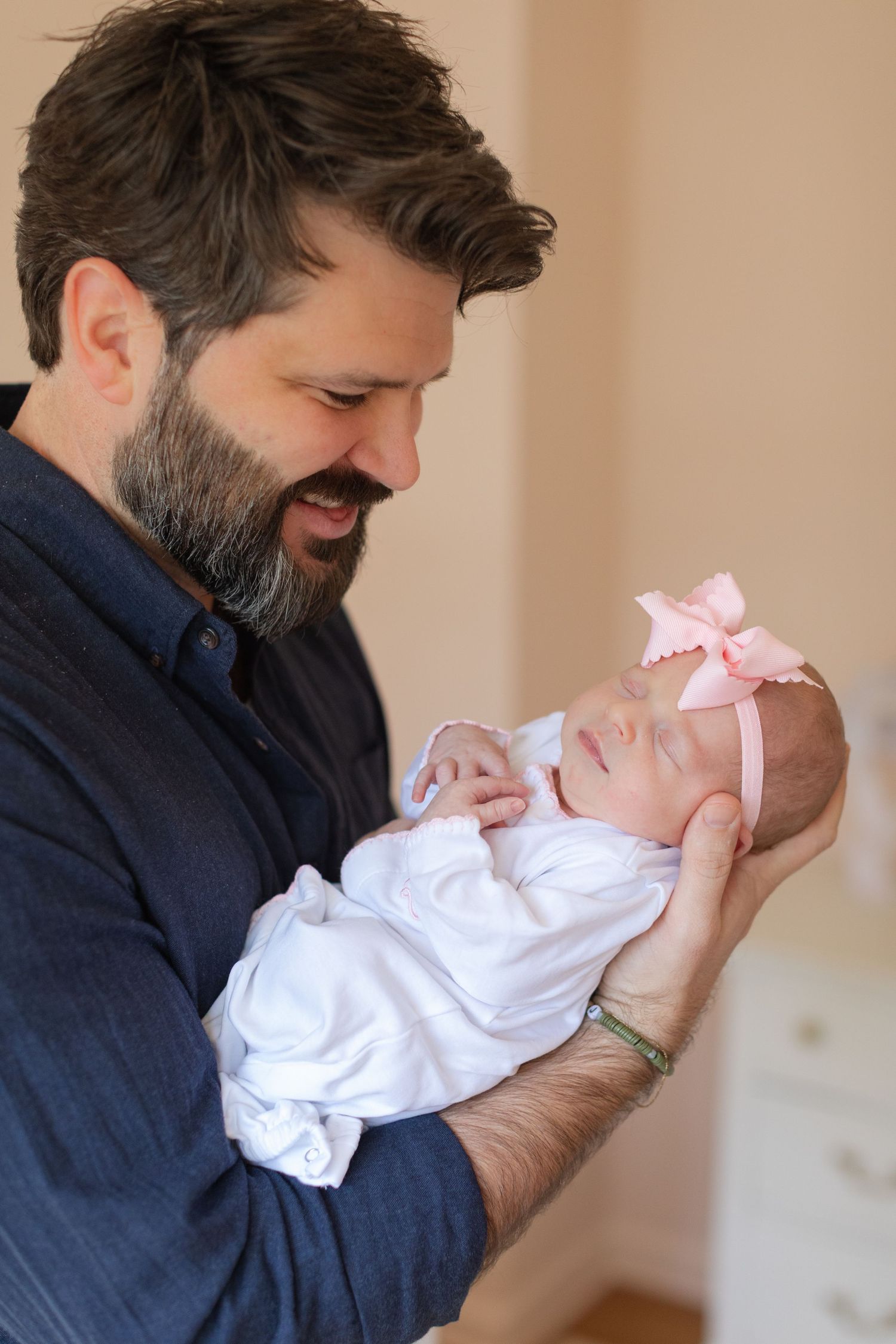A bearded parent in navy clothing holds a sleeping newborn wearing a pink bow.