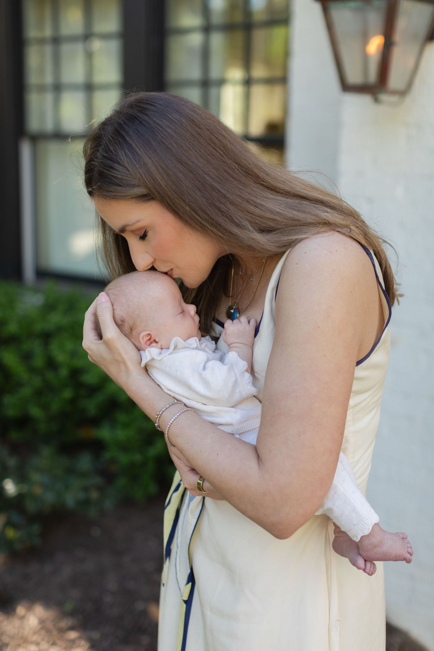 Young mother in yellow dress tenderly kissing her newborn baby outdoors near a white home entrance.