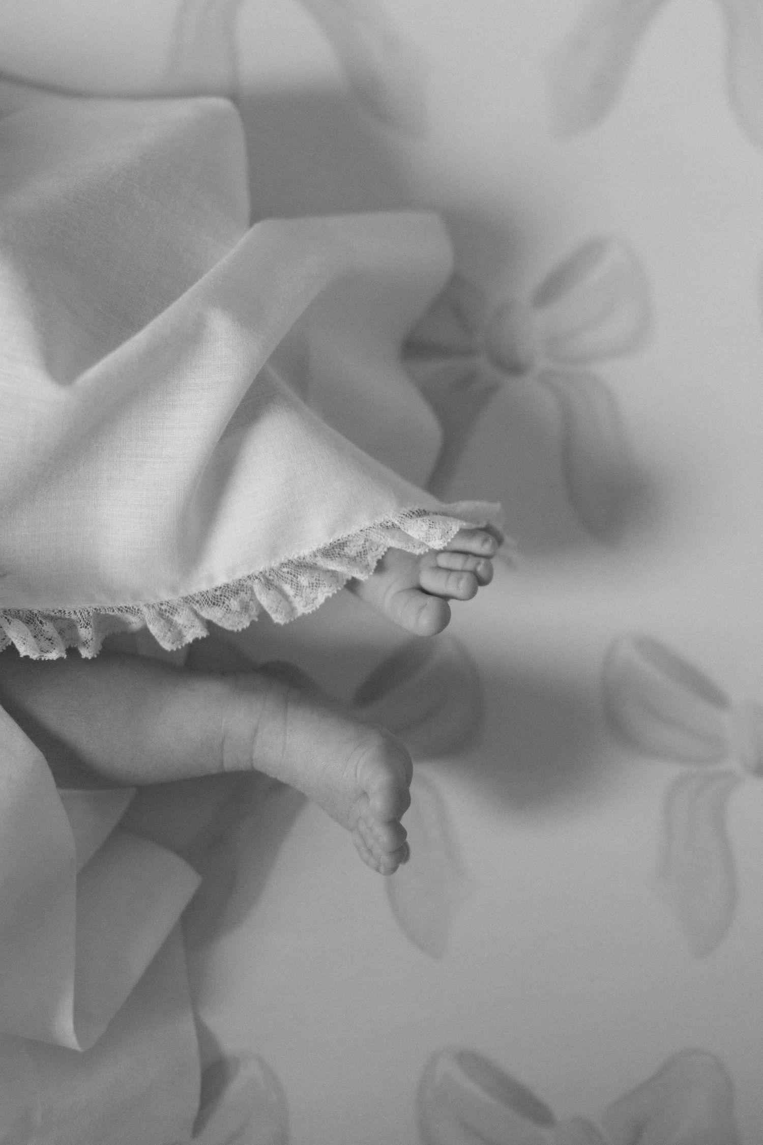 Black and white photo of tiny newborn baby feet peeking out from a delicate lace-trimmed blanket with bow pattern.