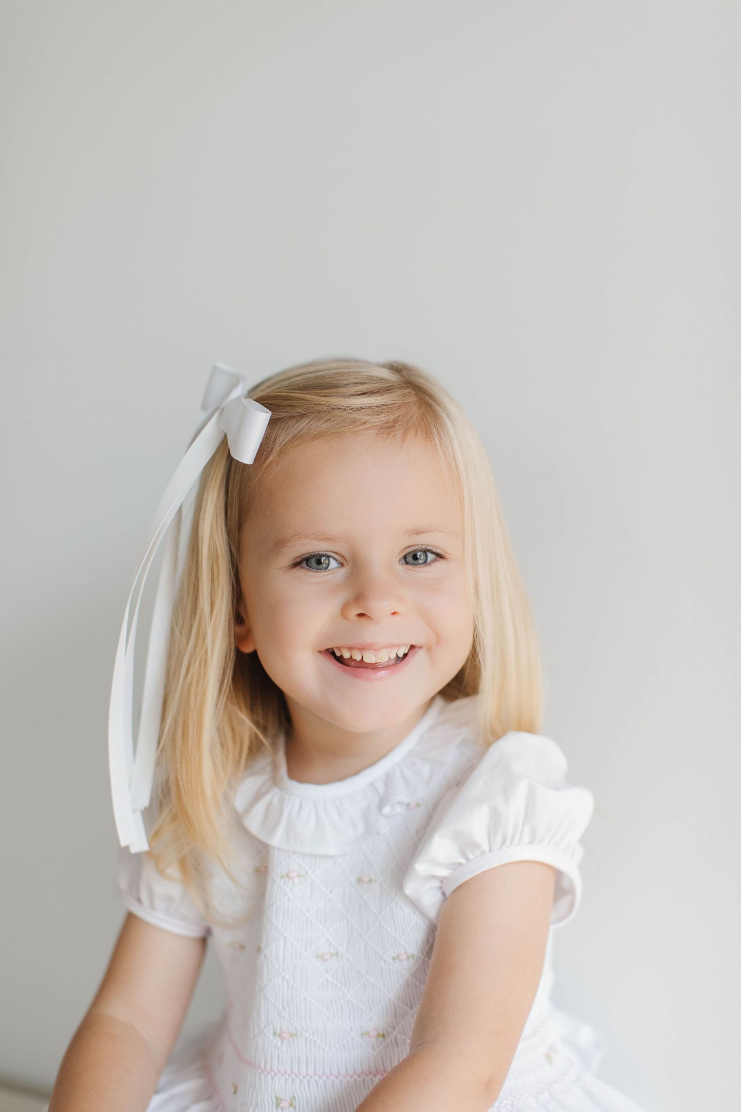 Child wearing white summer dress and ribbon hair bow smiles brightly against neutral background.