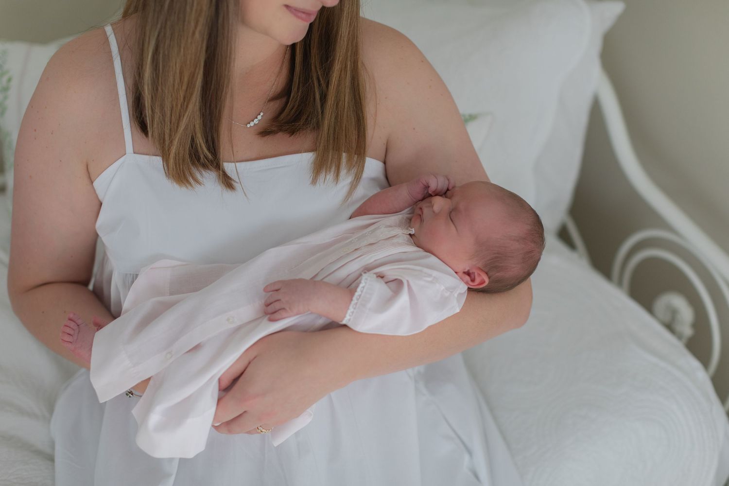 A sleeping newborn baby swaddled in white is cradled in someone's arms while resting on a white bed.