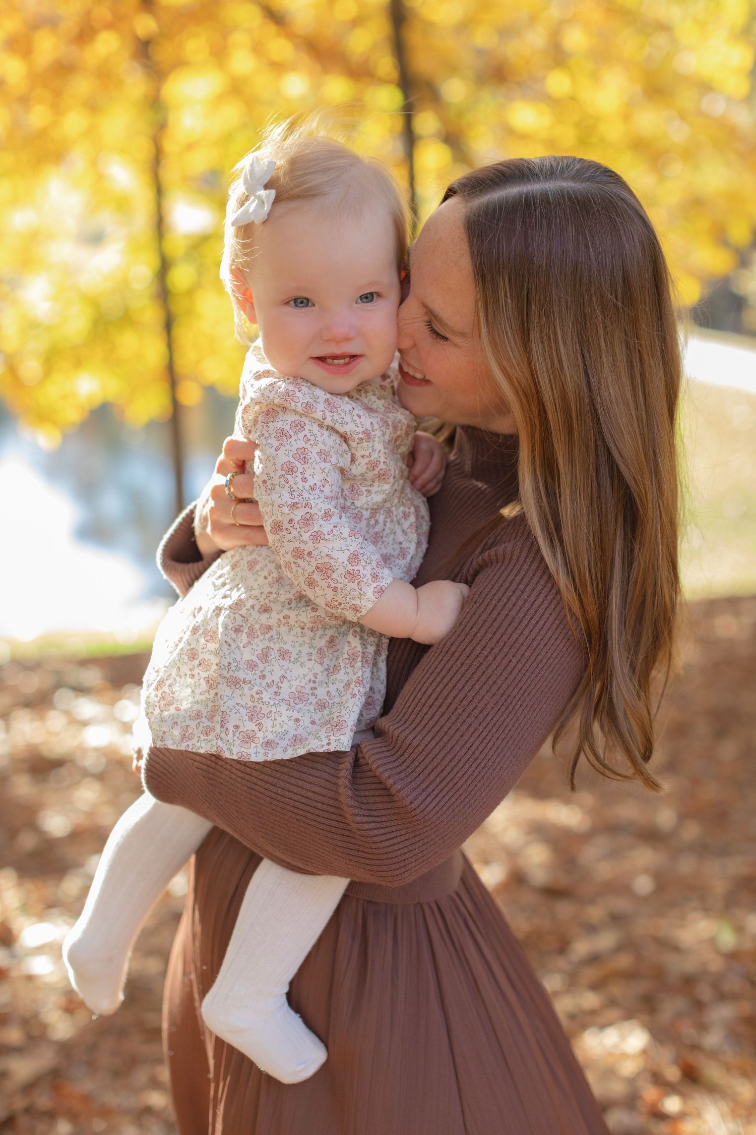 A tender moment between parent and baby against a backdrop of golden autumn leaves in a park setting.