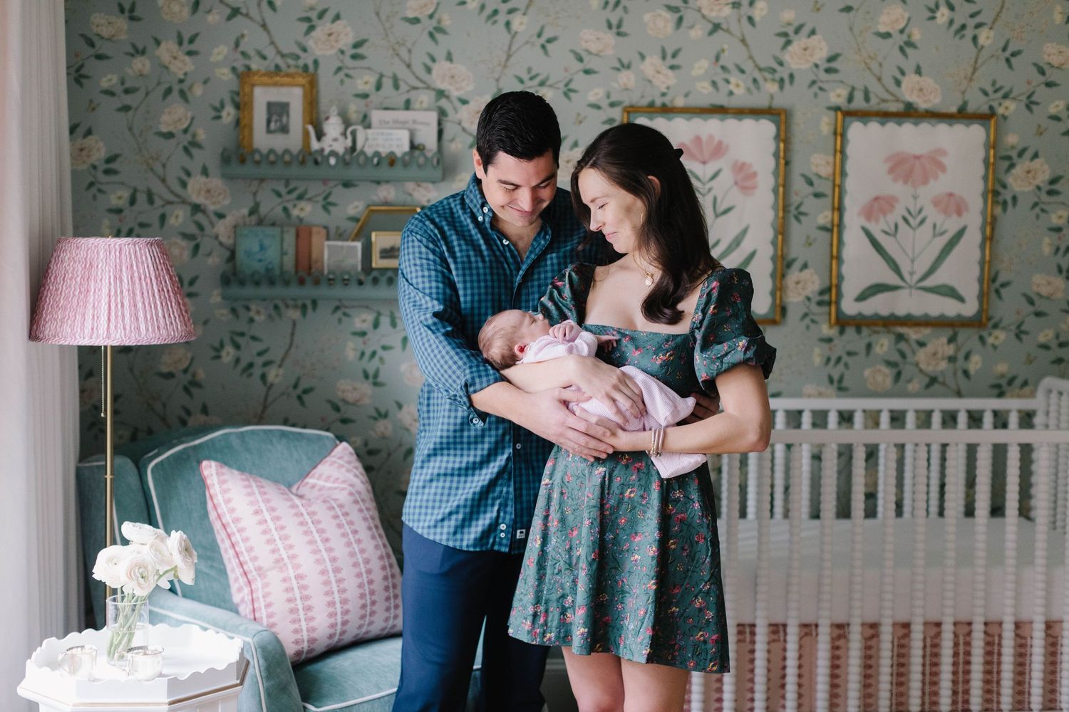 New parents admire their newborn baby in a nursery with floral wallpaper and pink decor.