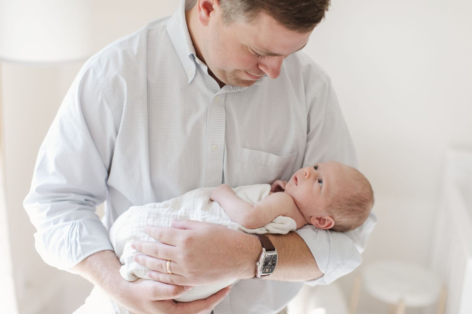 Parent in light blue shirt holds sleeping newborn against white background in minimalist setting.
