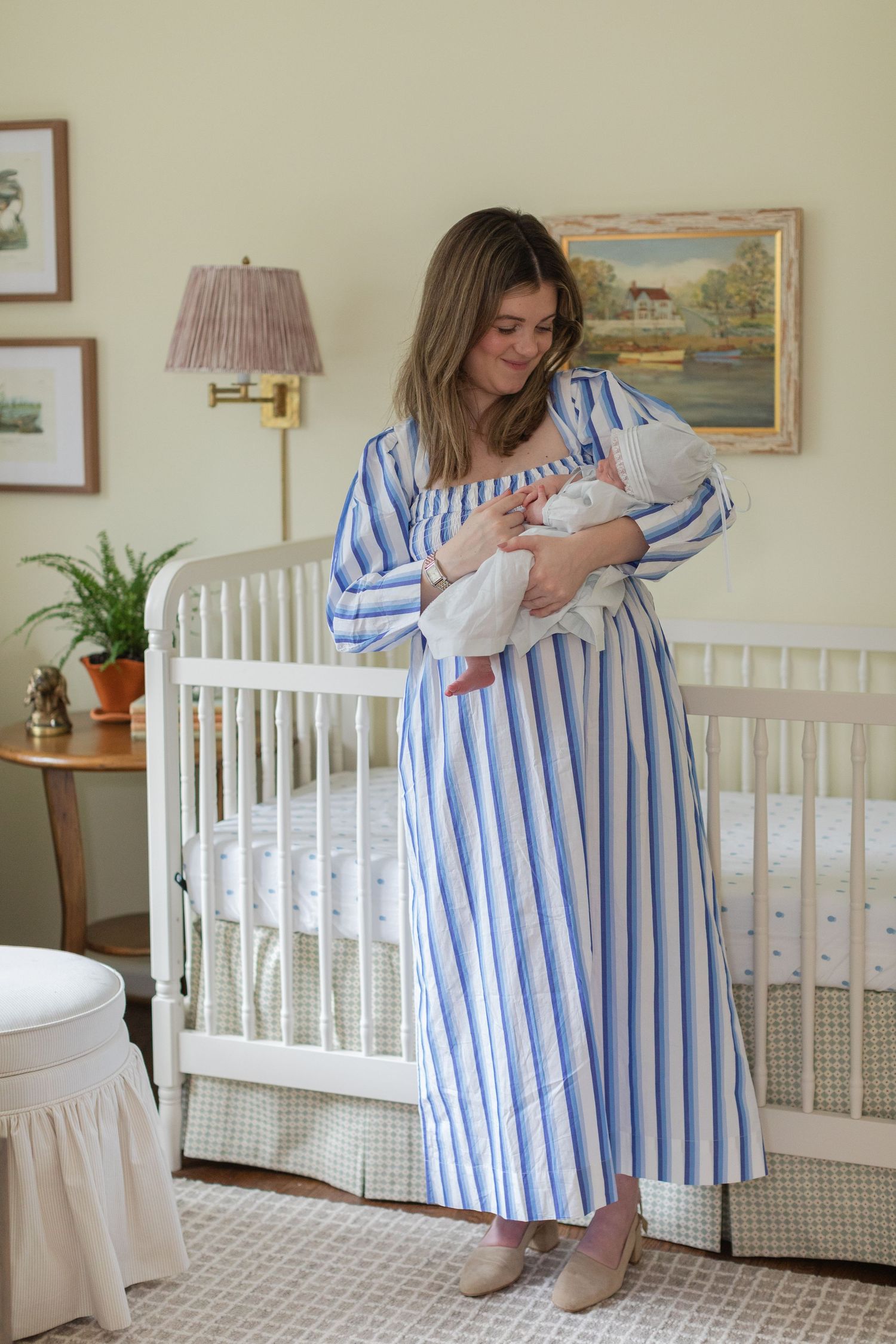 A person in a blue striped dress holds a newborn baby in a nursery with a white crib and framed artwork on the wall.