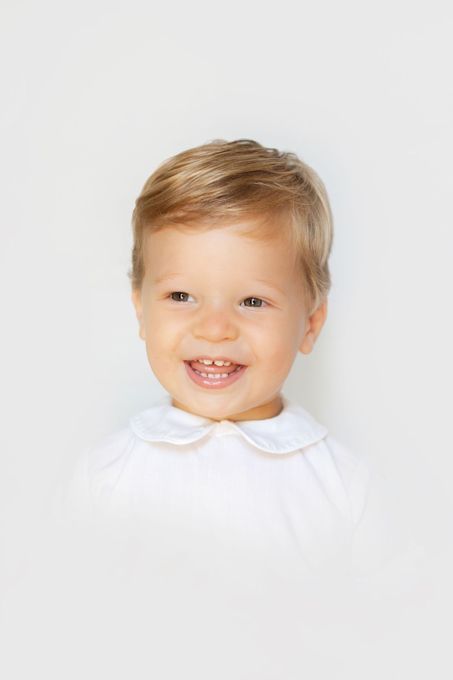 Adorable smiling toddler with blonde hair wearing white outfit against a clean white background.