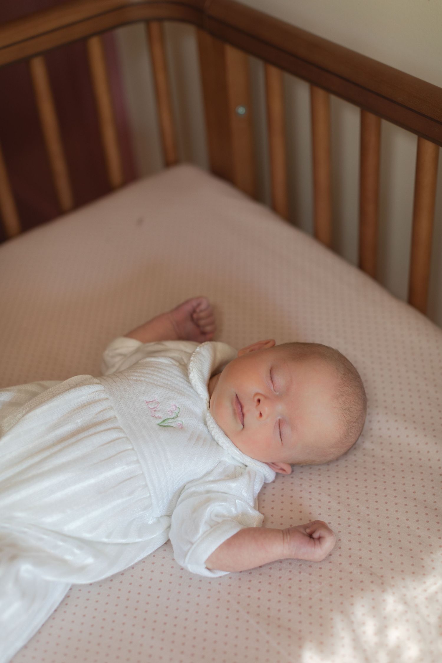 Newborn baby sleeping peacefully in a wooden crib, wearing a white dress on a light pink dotted mattress sheet.