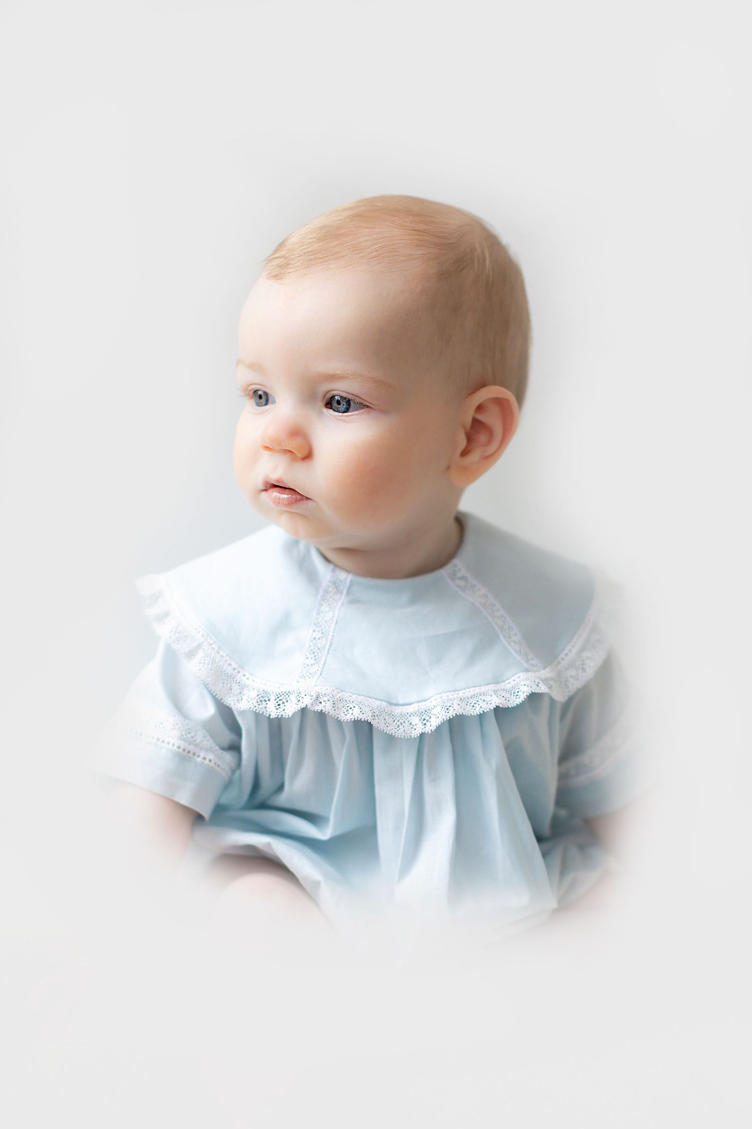 Adorable baby in light blue lace-trimmed dress gazing sideways with curious expression on white background.