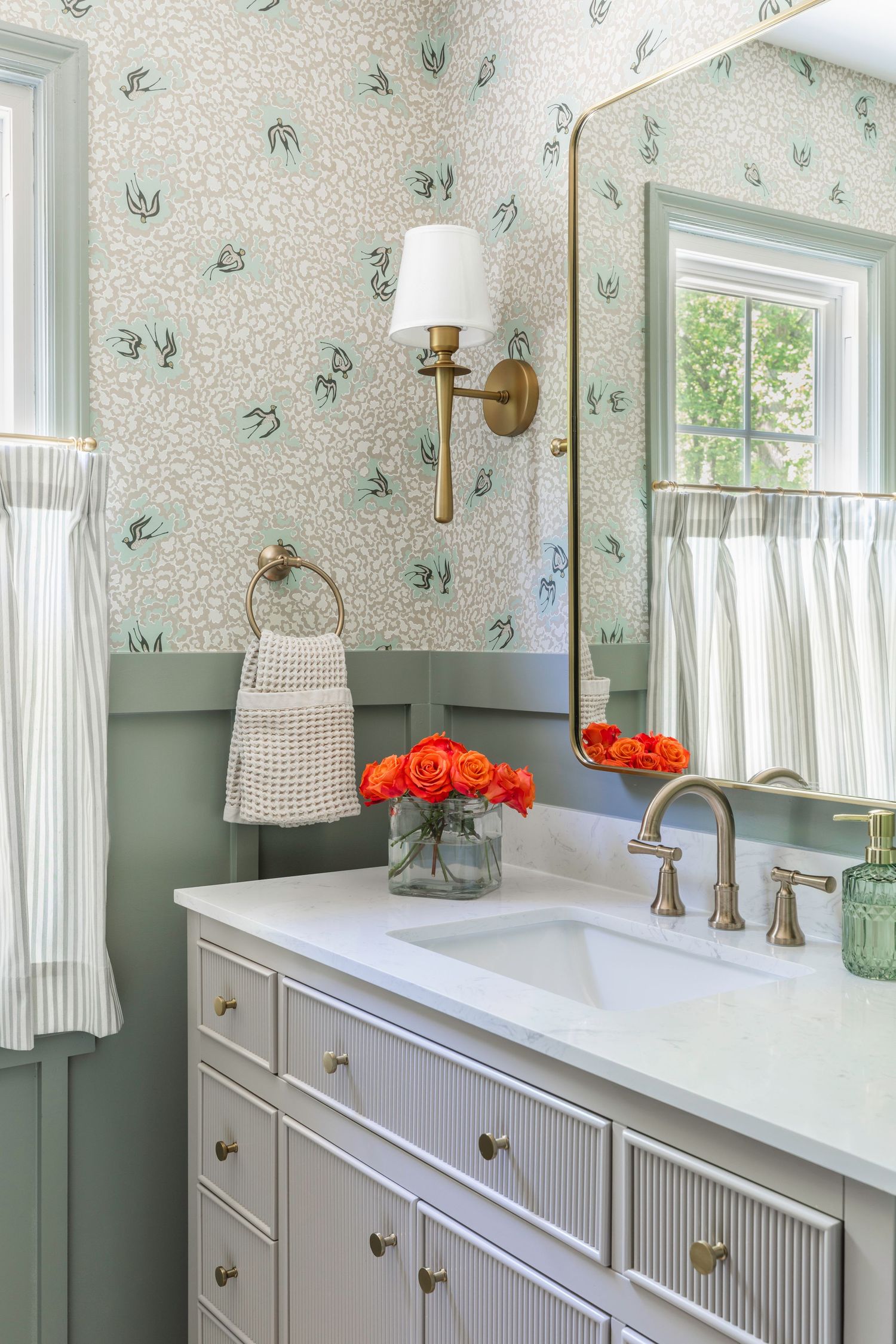 Elegant bathroom with floral wallpaper, gray vanity, brass fixtures, gold mirror, orange roses, and striped curtains.