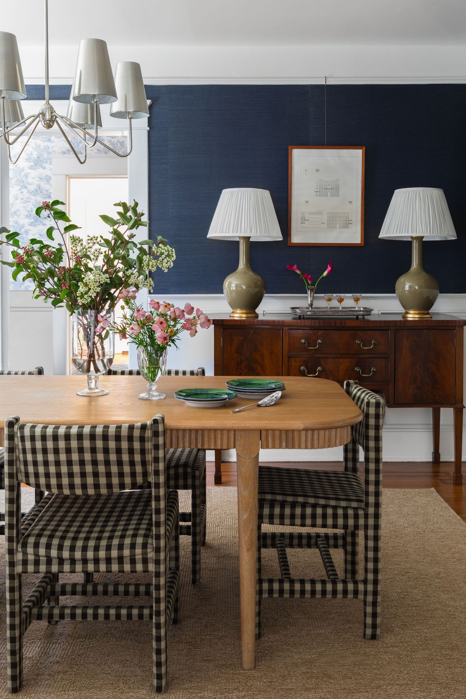 Navy blue dining room with gingham chairs, wood table, brass sideboard, pleated lamps, and floral centerpiece.