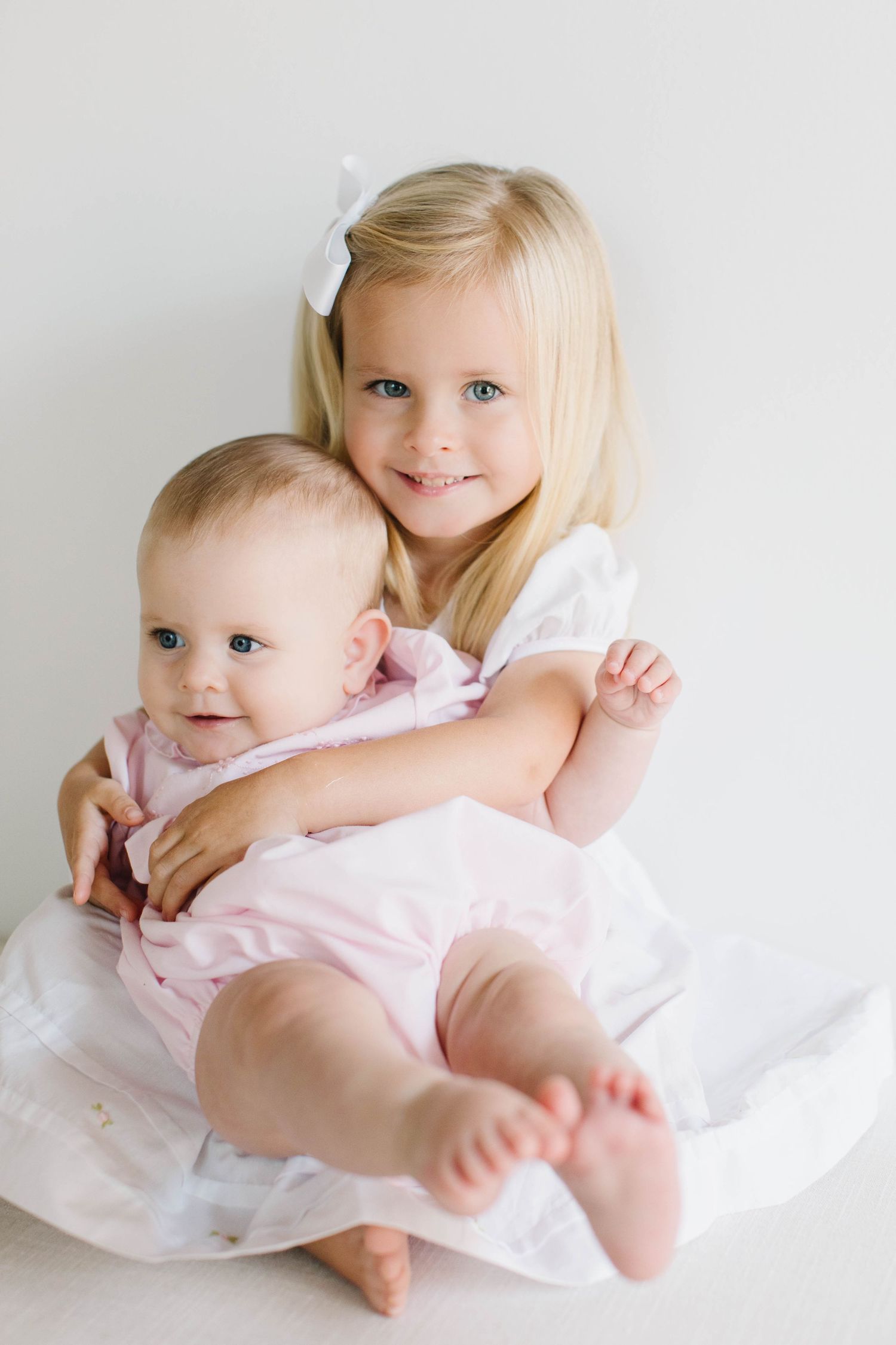 Children in white dresses sitting together against a minimalist backdrop showing sweet expressions and sisterly bond.