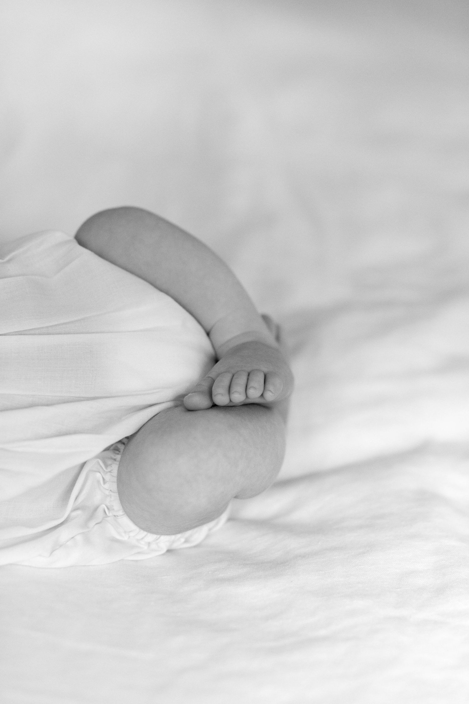 Close-up black and white photography of small baby feet against soft white bedding creating gentle shadows.
