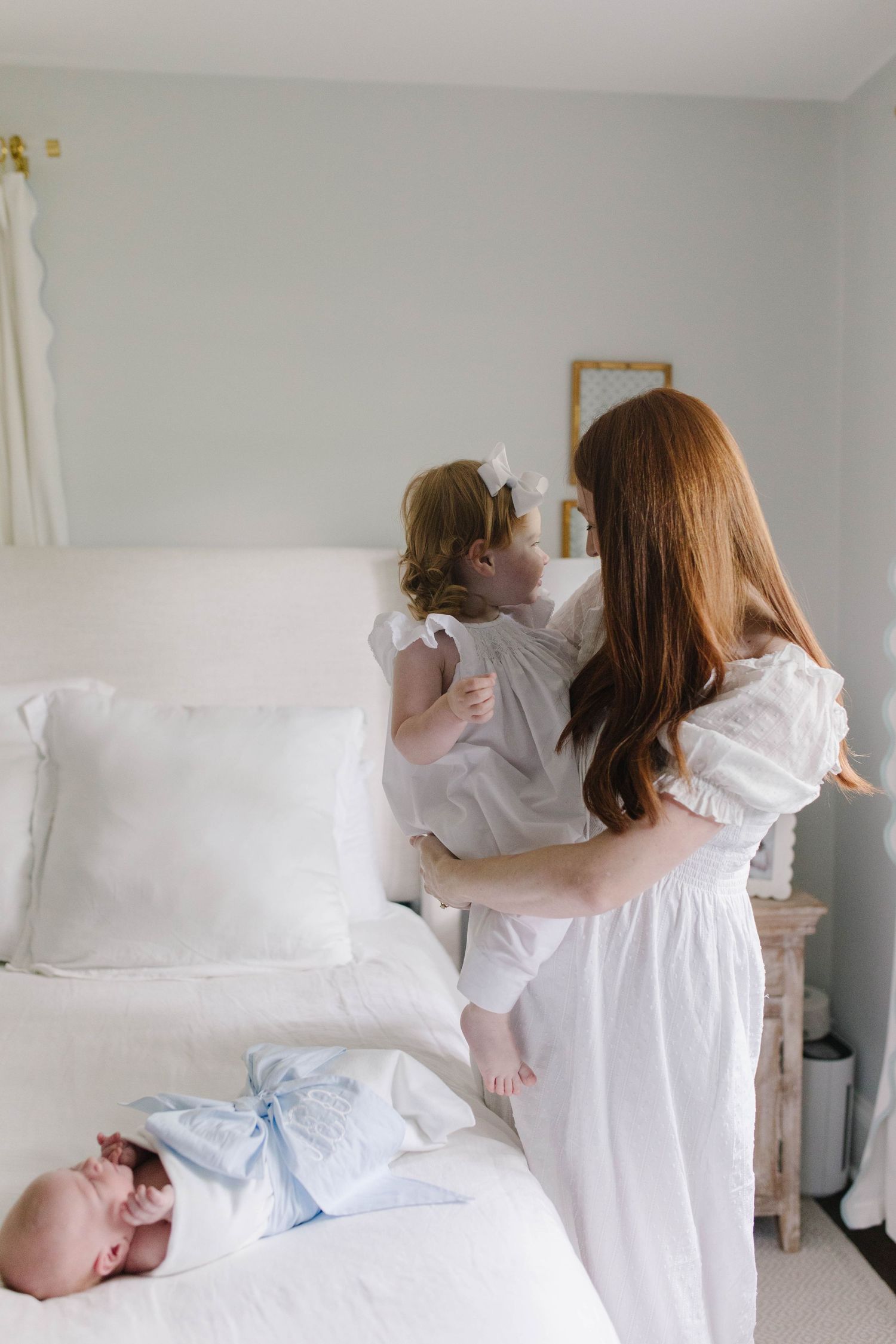 Children in white dresses playing together on white bed in bright bedroom.