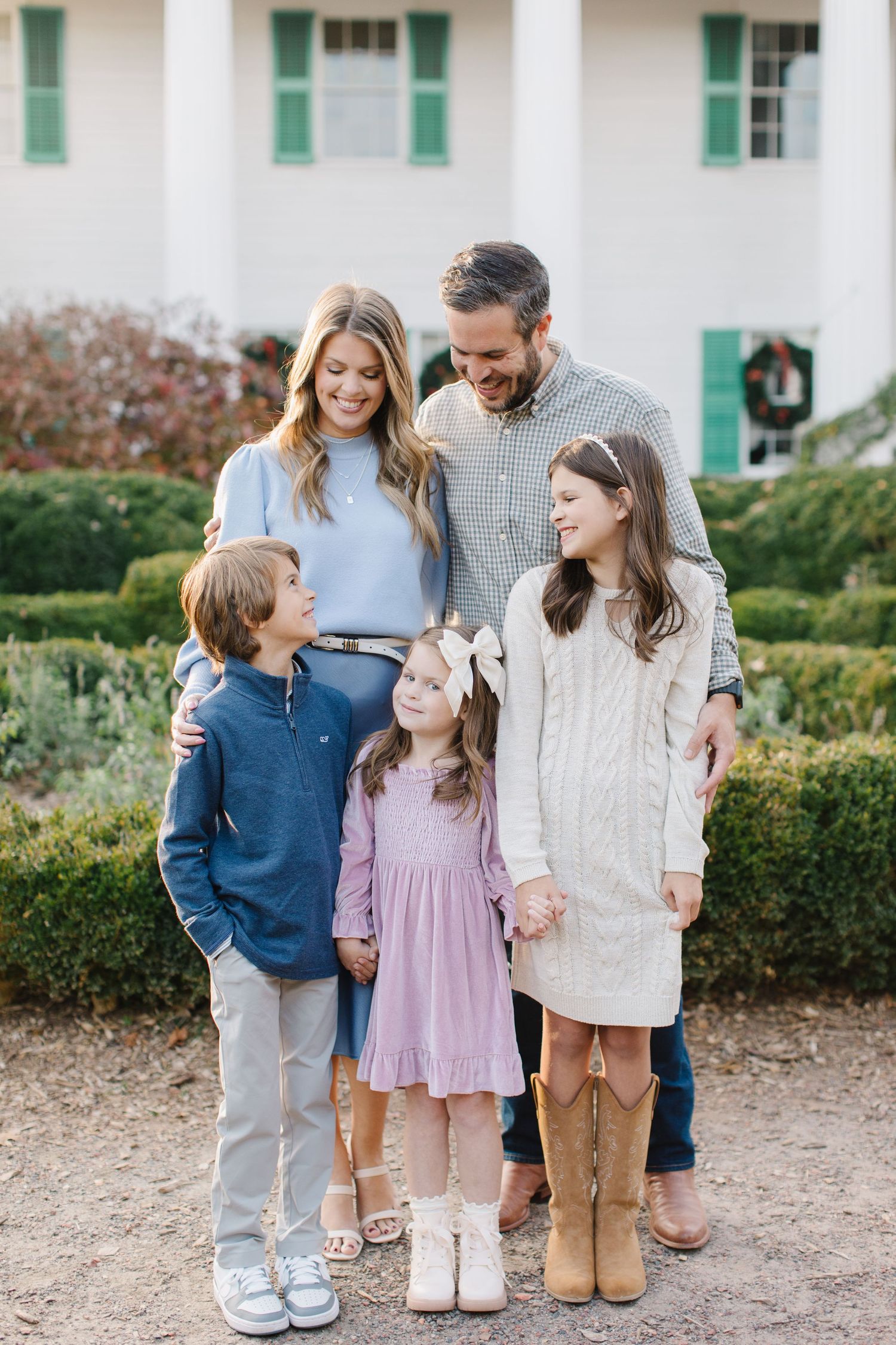 A family wearing coordinated casual outfits stands together on a gravel path near hedges and a white building.