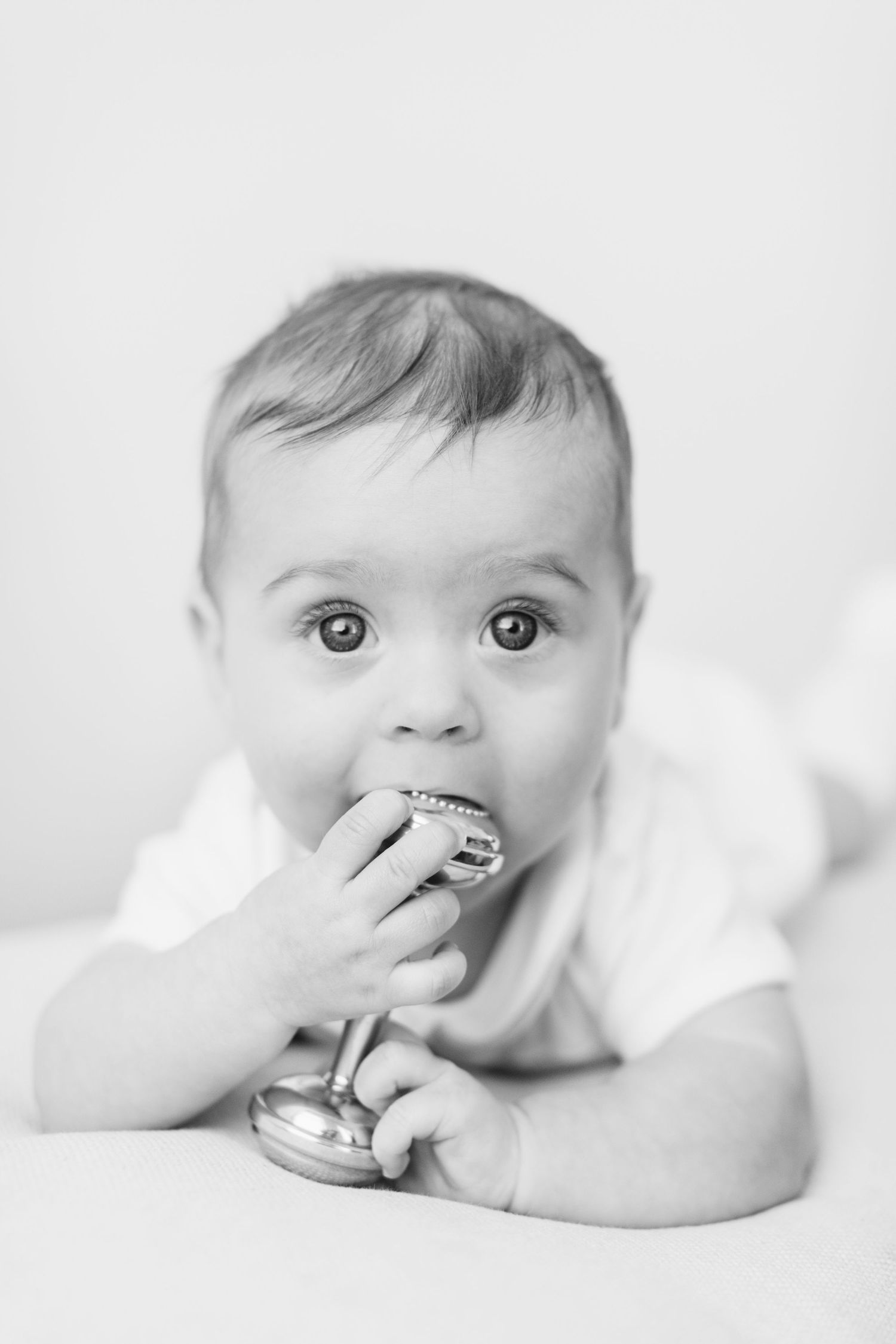 Black and white photo of an adorable baby chewing on a silver rattle while lying down.