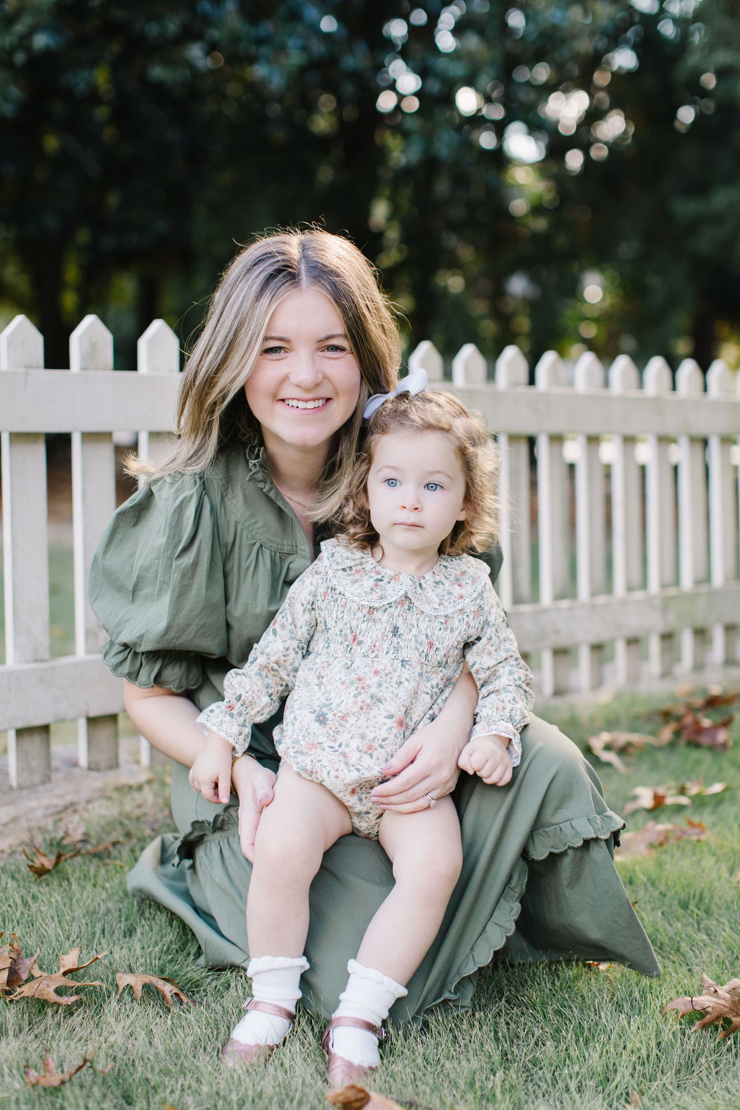 A parent and child sit together on green grass near a white picket fence during an outdoor photo session.