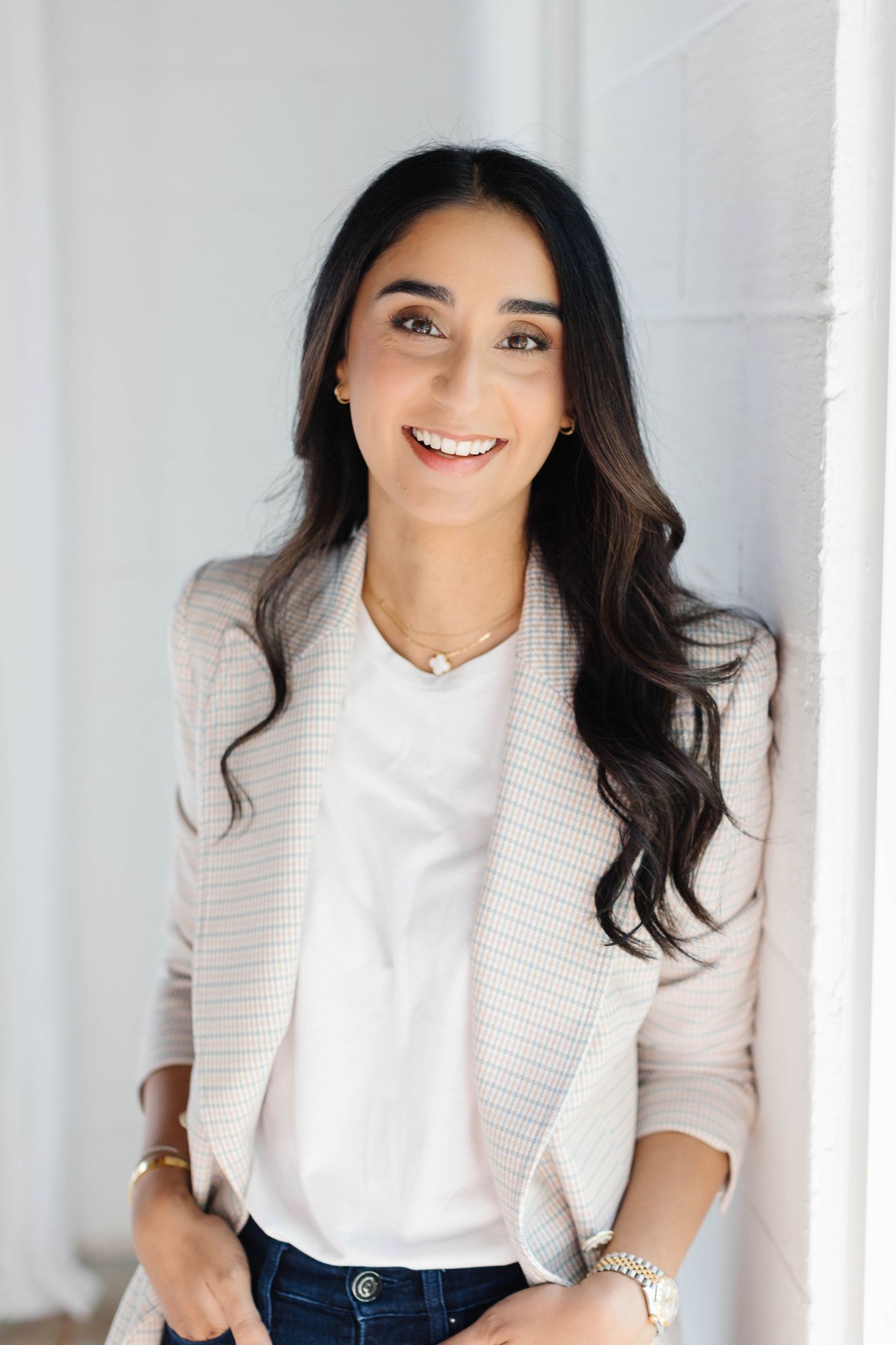 Professional woman in blazer smiling confidently, wearing a necklace and watch, leaning against a white wall.