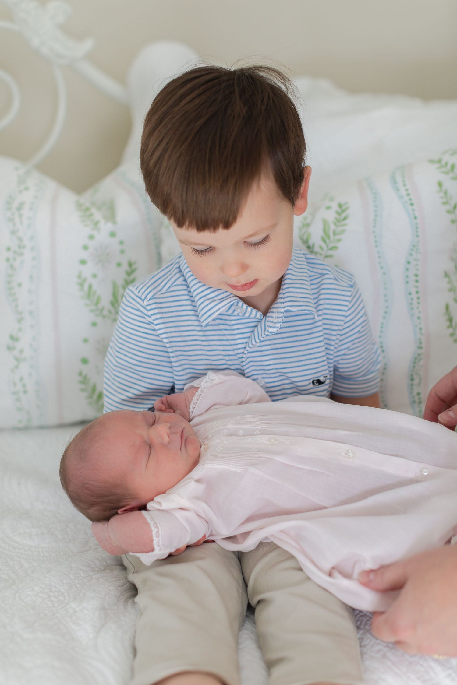A young child in a striped shirt gently interacts with a sleeping newborn baby in a pink outfit on a white couch.